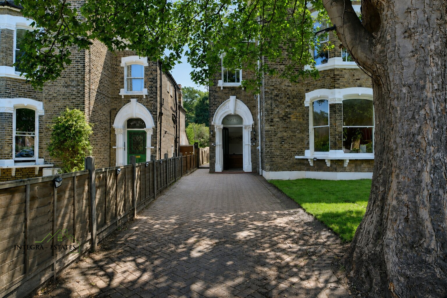 Residential street view with two adjacent brick houses featuring white trim around windows and doors. Left house has green door with arched white frame and number ‘324’ above, right house has similar arched entrance with number ‘327A.’ Paved driveway runs between houses toward back, wooden fence on left side. Large tree with thick trunk and green leaves provides shade, small grassy lawn visible in front of right-hand house. Logo ‘INTEGRA ESTATES’ in lower left corner.