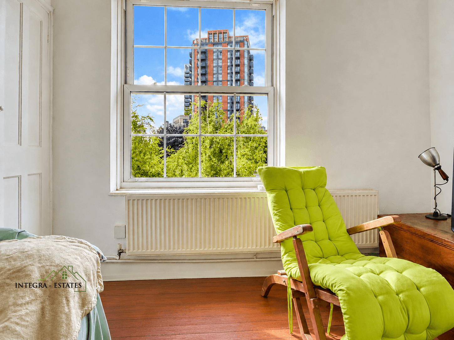 Bright cozy room with large window showing tall modern building surrounded by green trees under a blue sky. Interior features wooden floor, white radiator, wooden desk with metallic lamp, and wooden lounge chair with vivid lime-green cushion. Part of a bed or sofa with beige blanket printed ‘INTEGRA ESTATES’ and house logo visible.