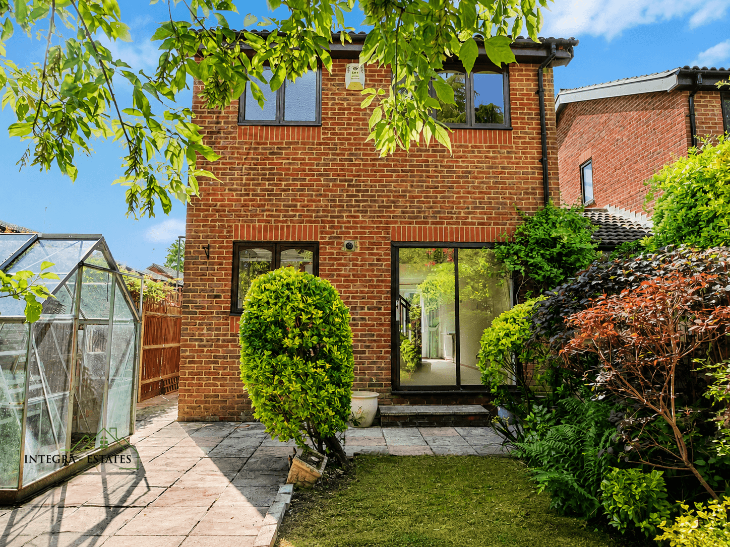 Rear exterior of two-story brick house with small well-maintained garden. Upper floor has two windows, ground floor features large sliding glass door opening to stone-paved patio bordered by shrubs and plants. Glass greenhouse structure visible to left beside wooden fence, colorful foliage to right. Bright sunny day with leafy tree branches framing top. Watermark ‘INTEGRAL ESTATES’ in lower left corner.
