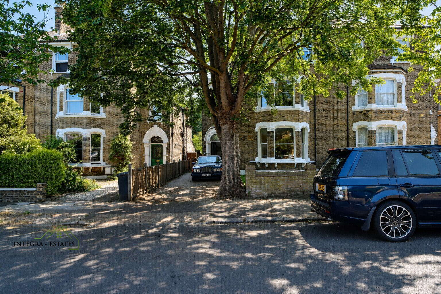 Residential street with two large brick houses featuring white trim and bay windows, traditional Victorian or Edwardian style. Tall tree in center foreground provides shade, driveway between houses with silver car parked, dark blue SUV along curb to right. Sunlight filters through leaves creating pleasant suburban atmosphere. Logo ‘INTEGRA ESTATES’ in lower left corner.