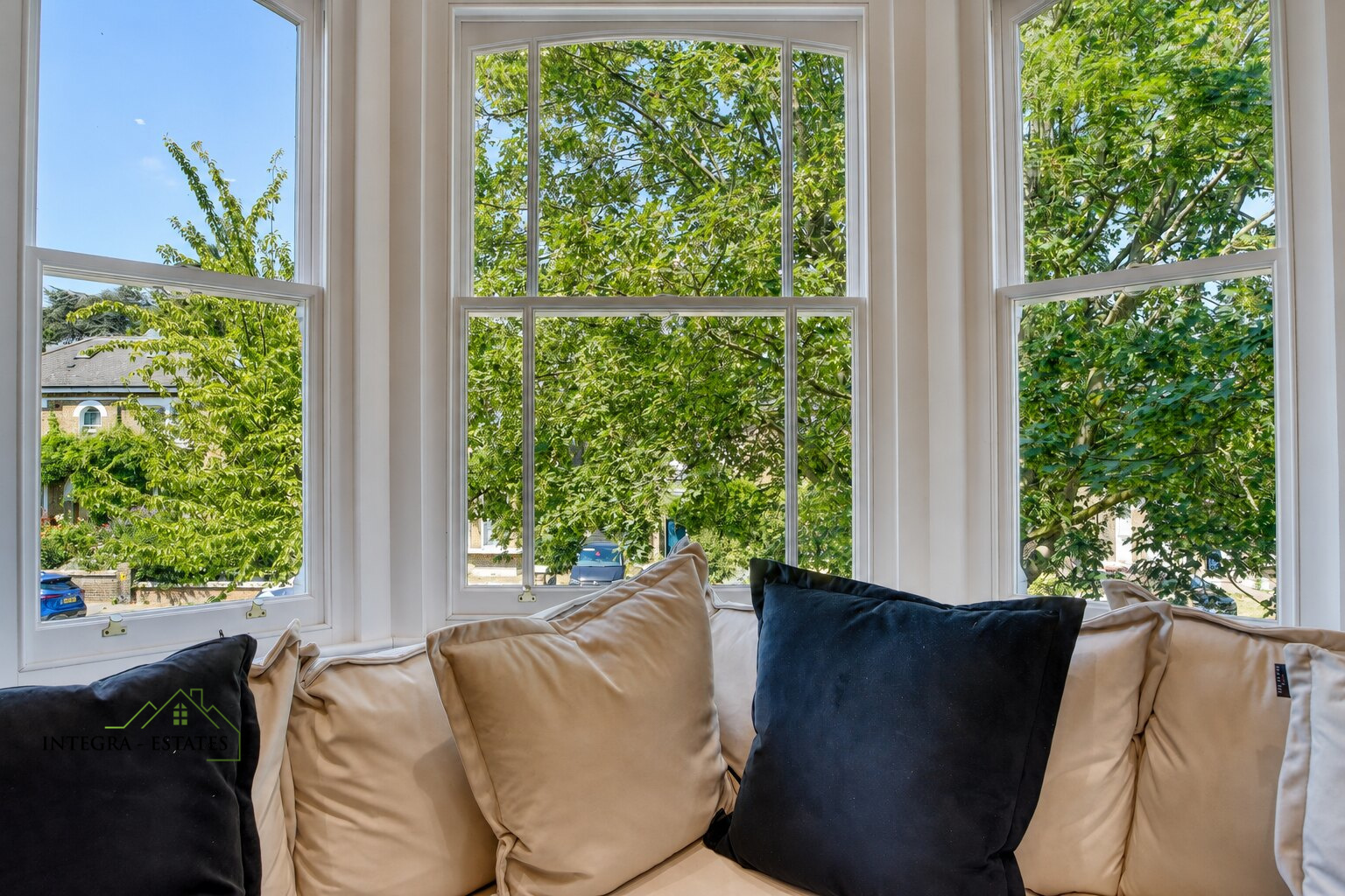 Cozy indoor seating area with beige cushioned bench in front of large bay window, decorated with beige and dark blue pillows. Outside view shows lush green trees, residential street with houses and parked cars under sunny sky.