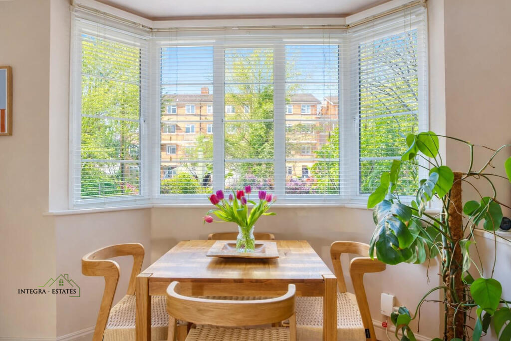 A bright dining area with a large bay window fitted with white blinds, allowing natural light to stream in. A wooden dining table with four woven‑seat chairs sits in front of the window, topped with a glass vase of pink tulips. A tall green houseplant with broad leaves stands to the right, while light‑colored walls and warm tones create an inviting atmosphere.