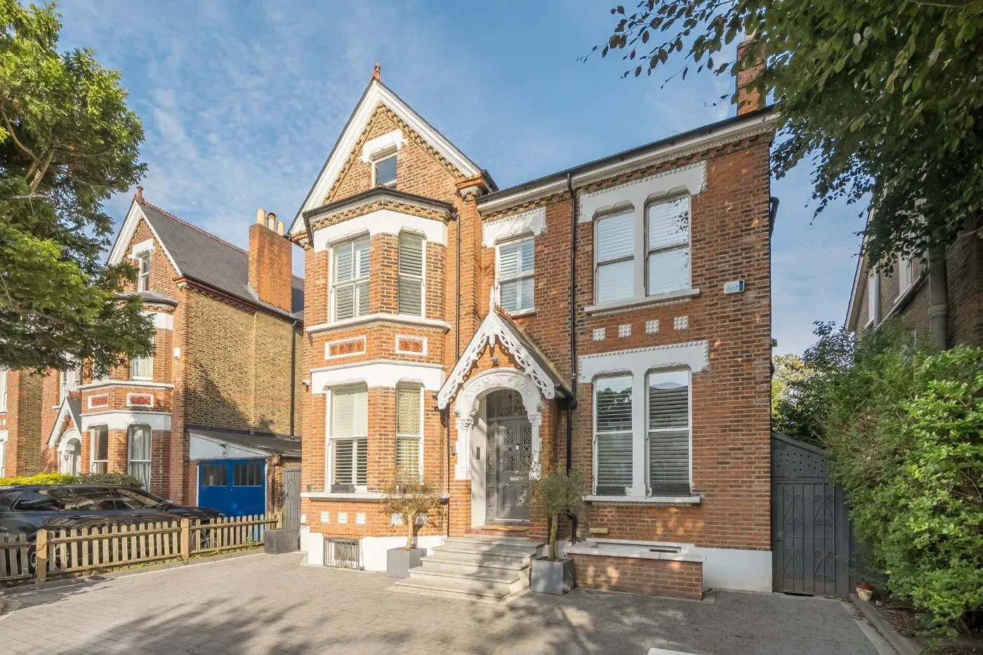 Two-story Victorian-style brick house with steeply pitched roof, bay windows, and ornate white trim. A gabled porch with decorative woodwork frames the front entrance, flanked by two small trees in square planters. The driveway is paved with light-colored bricks, and a wooden fence separates the property from a neighboring house with similar architecture