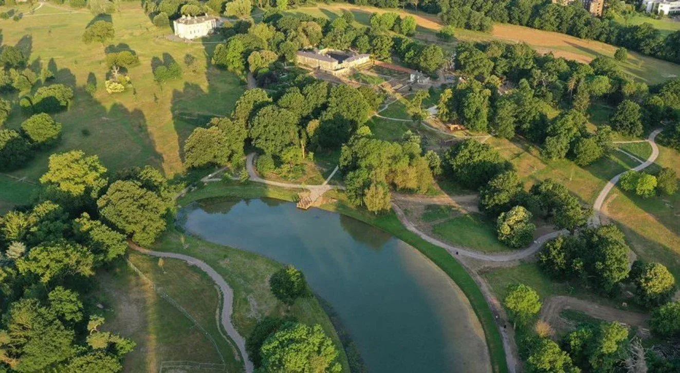 Aerial view of a landscaped estate featuring a curved pond with a small wooden bridge, surrounded by winding walking paths, grassy areas, and clusters of trees. In the upper section, several buildings are visible, including a large white structure and a historic-looking mansion. The scene is lush and thoughtfully designed, blending natural beauty with architectural elements.