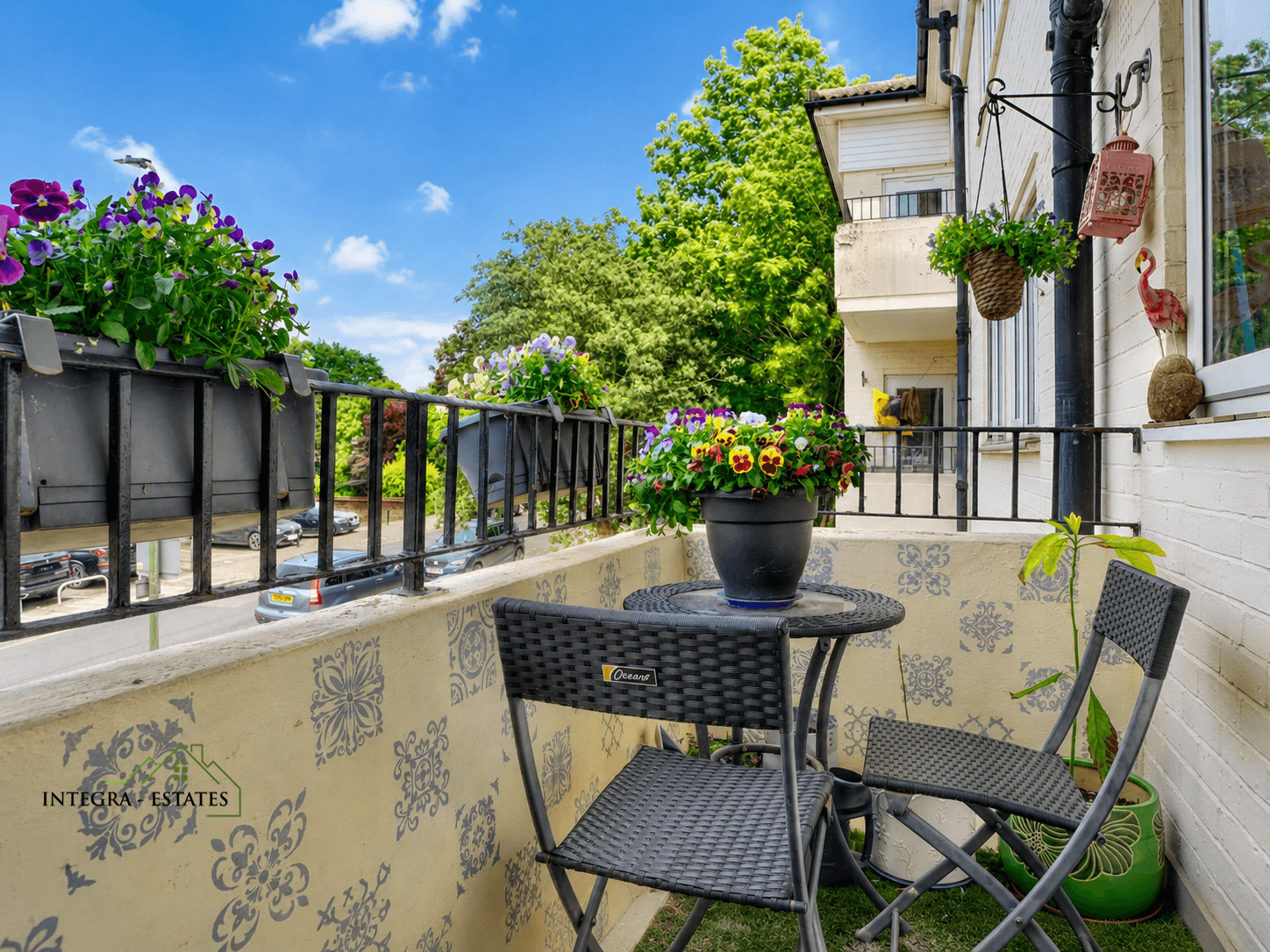 “Small charming balcony attached to light-colored building, decorated with patterned tile wall and black metal railing holding flower boxes of vibrant purple and yellow pansies. Round woven table with two matching chairs in center, black pot of colorful pansies on table. Hanging basket with greenery, red lantern, and decorative flamingo on wall near window. Background shows neighboring balconies, lush green trees, and parked cars below.