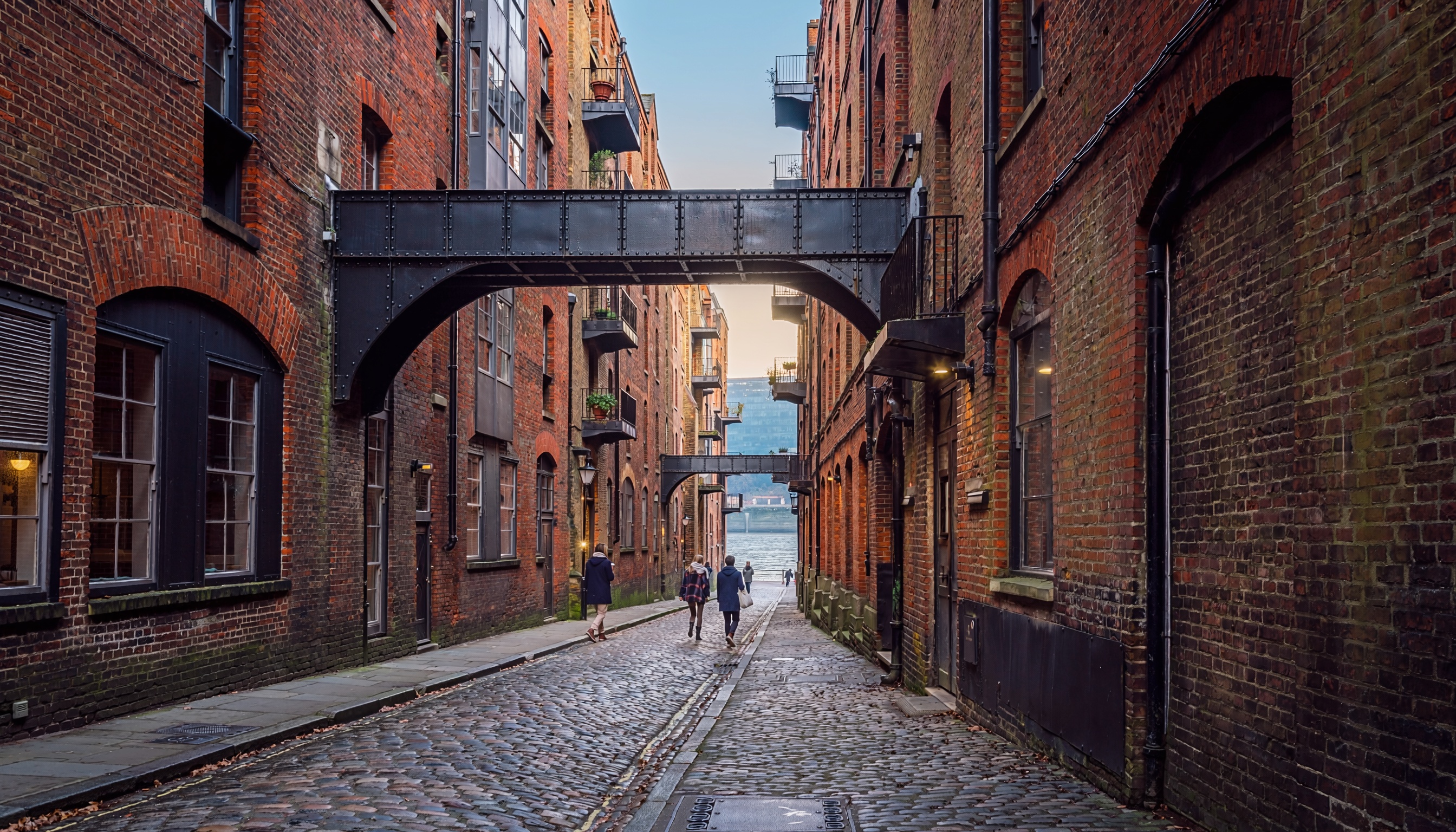 Narrow cobblestone street with tall brick buildings connected by black metal bridges, people walking toward distant waterfront.