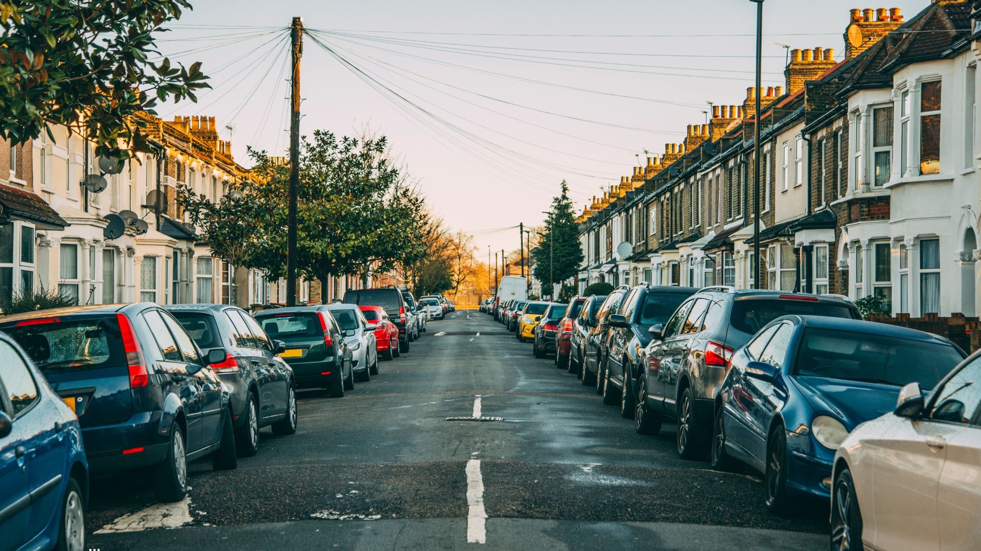 A residential street lined with parked cars on both sides, bordered by rows of traditional terraced houses. Overhead utility lines stretch across the road, and the scene is lit by warm afternoon sunlight, giving a typical suburban or South East London neighbourhood feel.