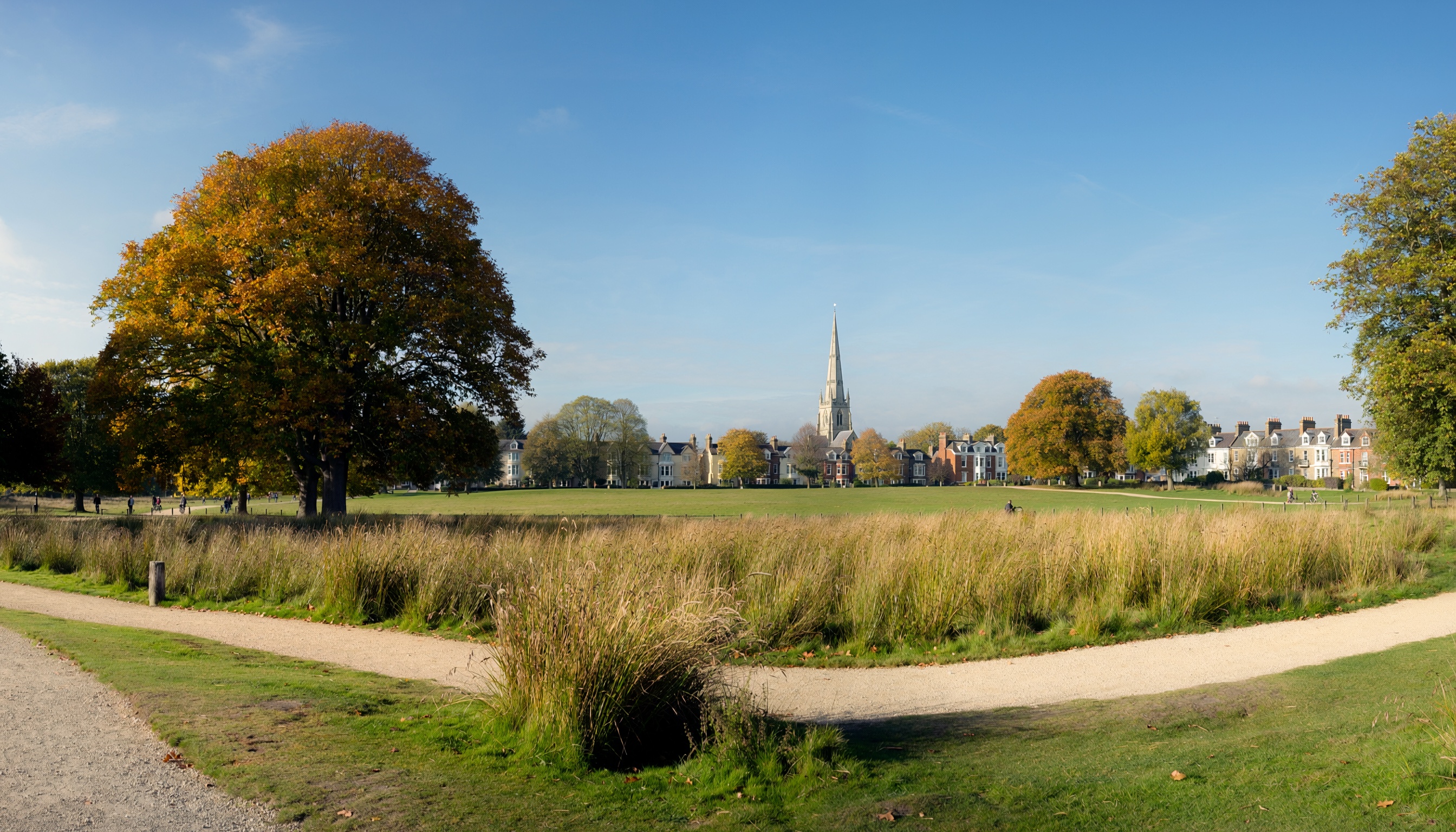 Open green park with gravel path, autumn trees, and a tall church spire rising behind traditional houses under a blue sky.