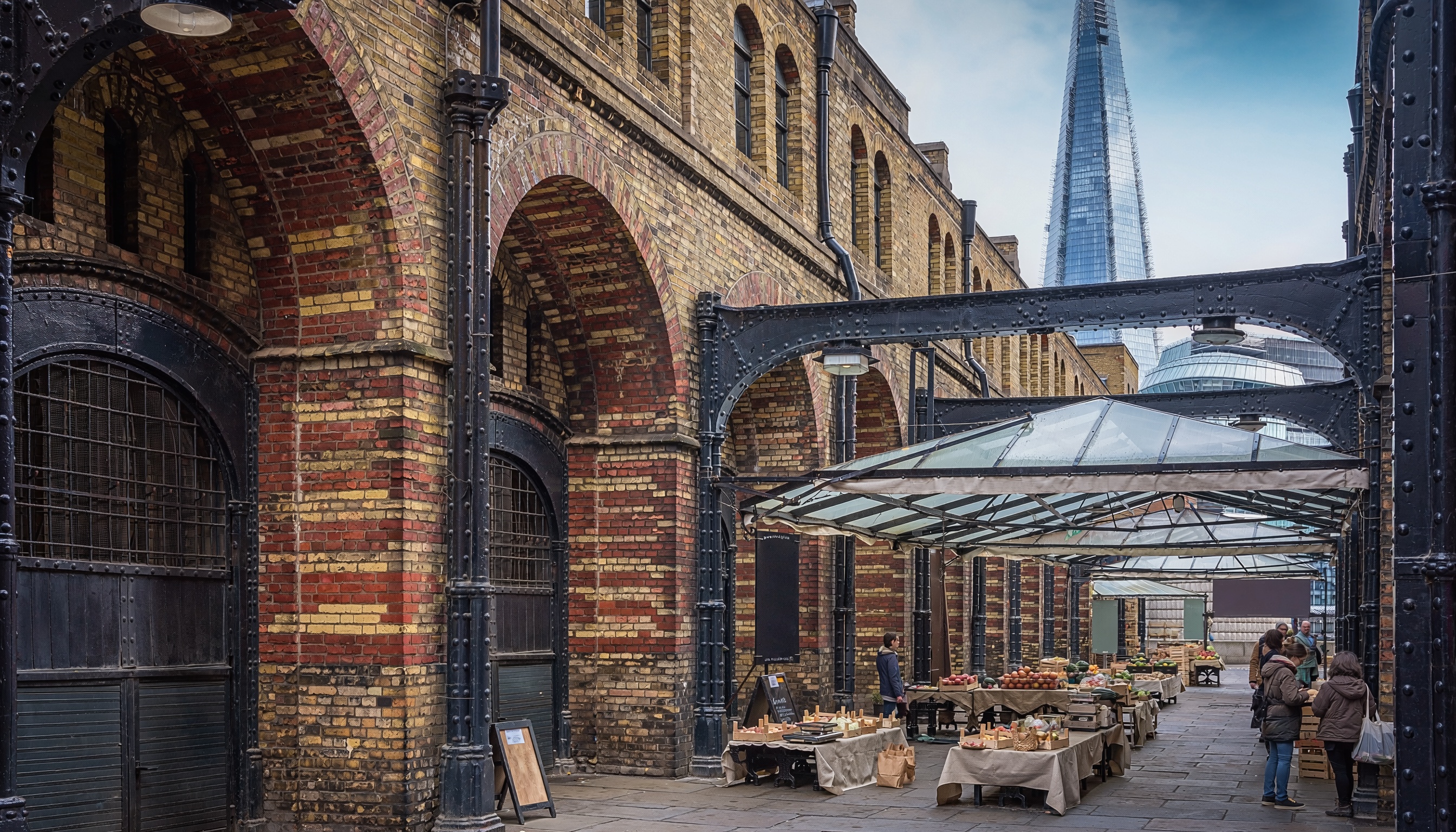 Outdoor market beneath glass canopy with stalls of fruits and vegetables, historic brick buildings, and The Shard skyscraper rising in the background.