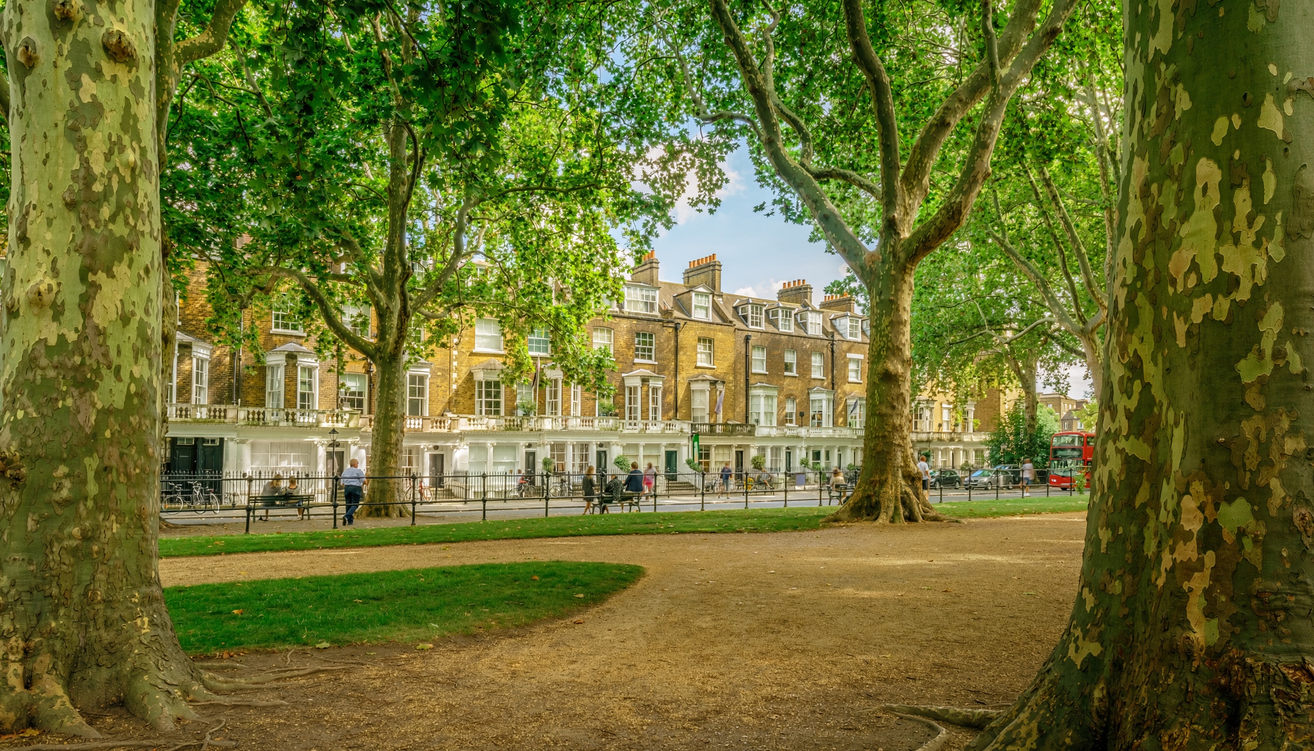 Urban park framed by large trees with Georgian-style townhouses in the background, people walking and sitting on benches, bicycles nearby, and a red double-decker bus partially visible.