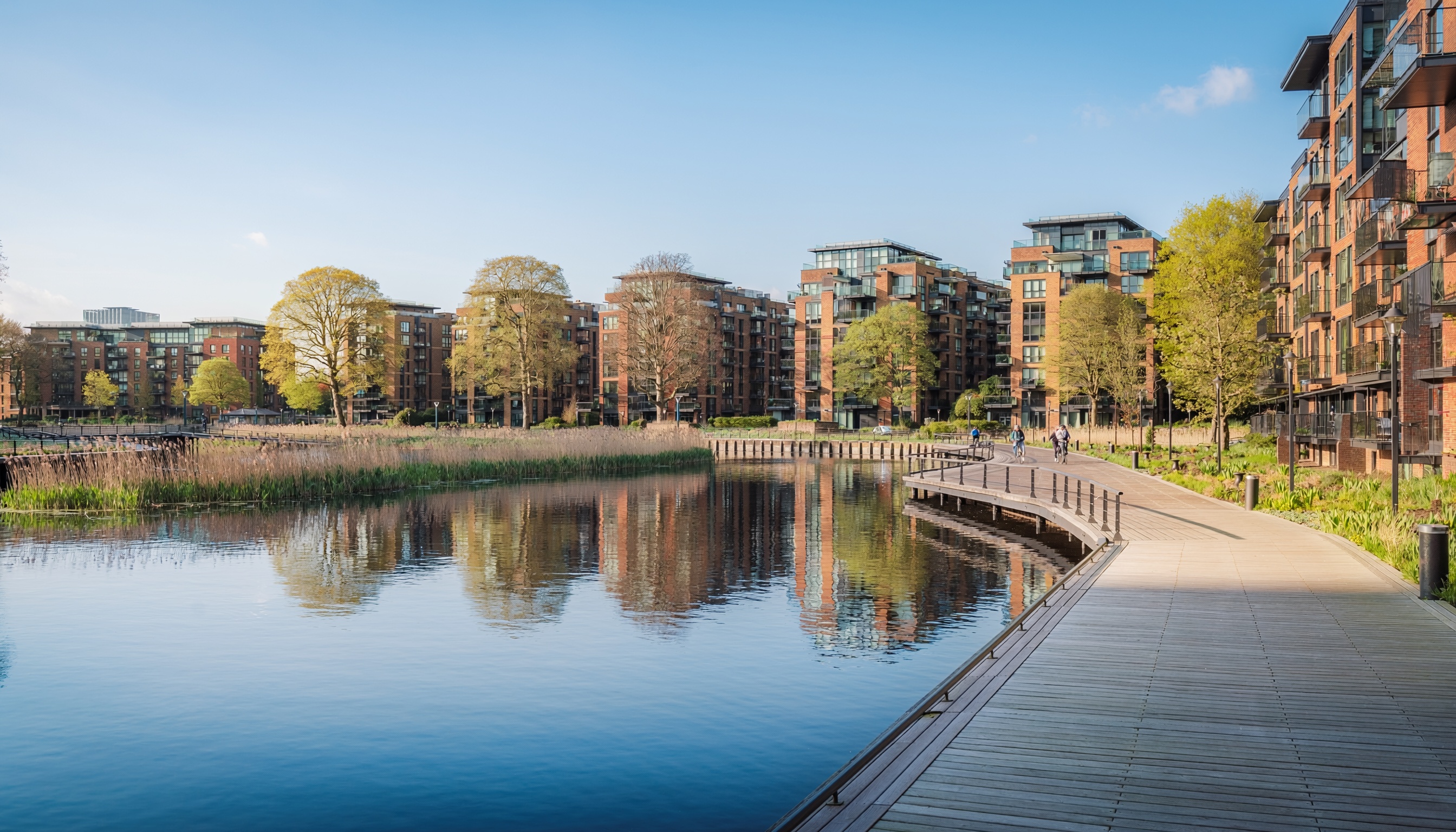 Urban waterfront with modern brick-and-glass residential buildings, trees with fresh green foliage, calm water reflecting the scene, and people walking or cycling along a wooden boardwalk.