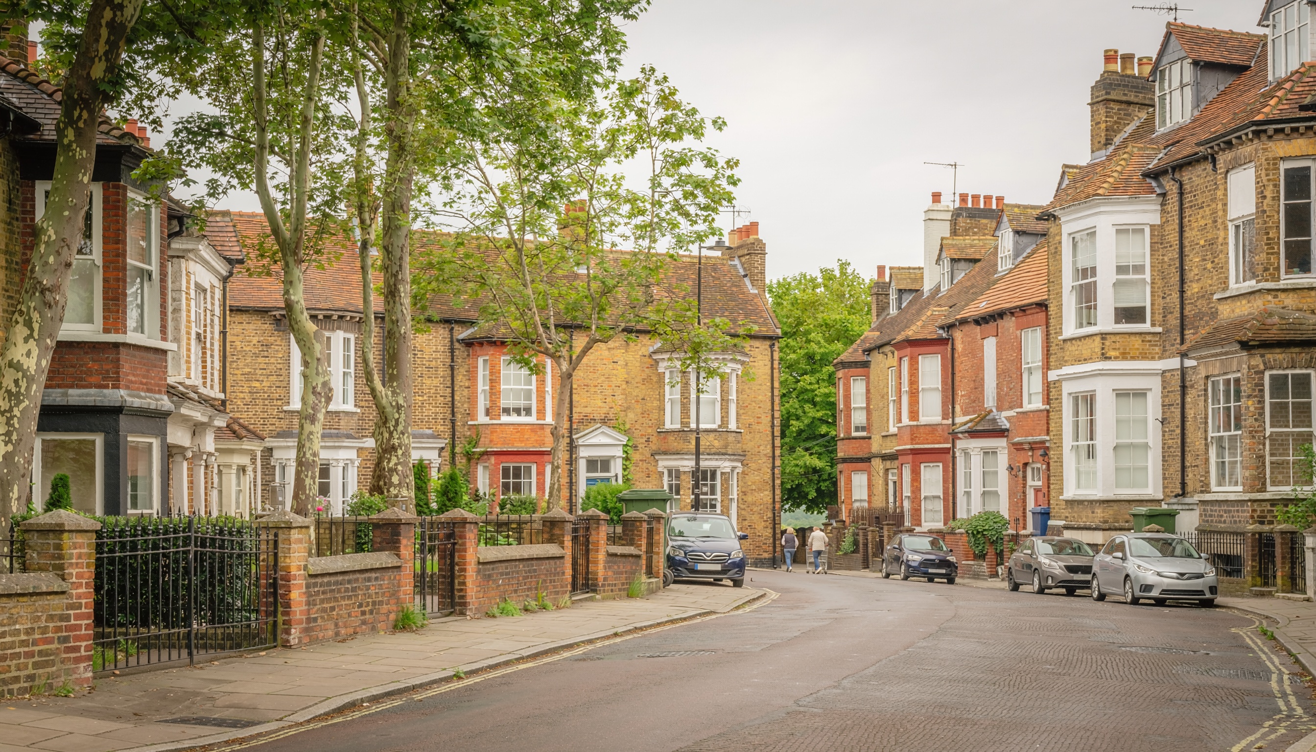 Residential street with traditional brick houses, bay windows, tiled roofs, small front gardens, parked cars, and tall trees lining the sidewalk.