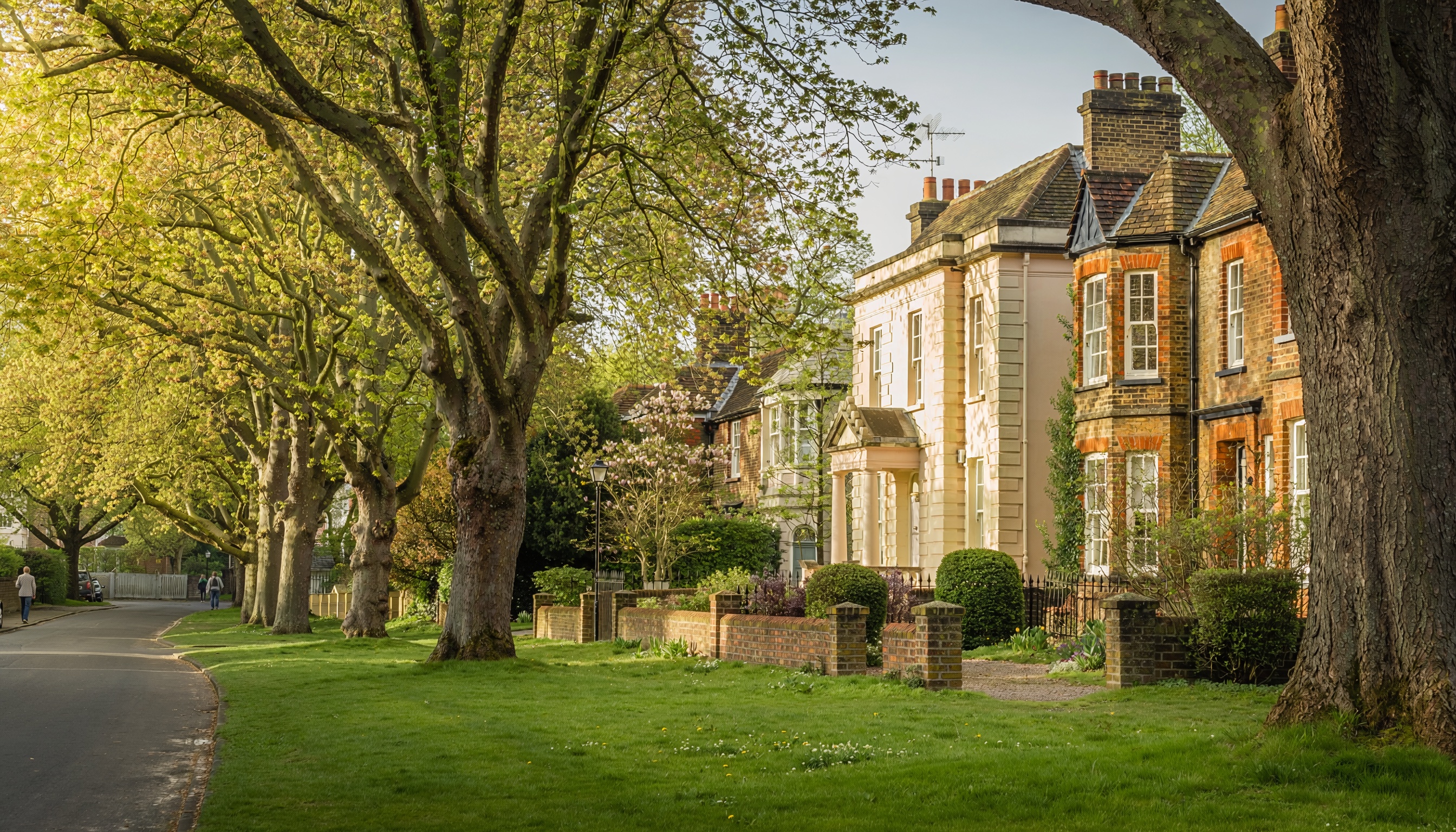 Curving residential street lined with mature trees and traditional brick houses, bay windows and chimneys, with warm sunlight filtering through in late afternoon.