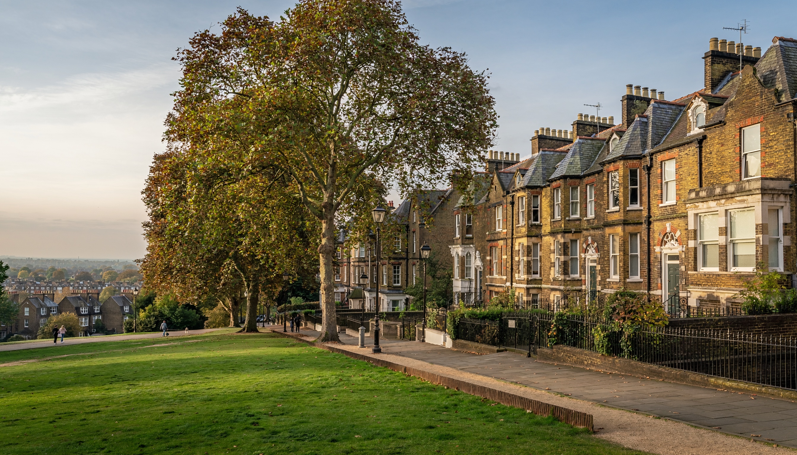 Victorian-style brick townhouses with ornate trim and slate roofs, fronted by iron railings and street lamps, beside a grassy park with mature trees and people walking along the paved path.