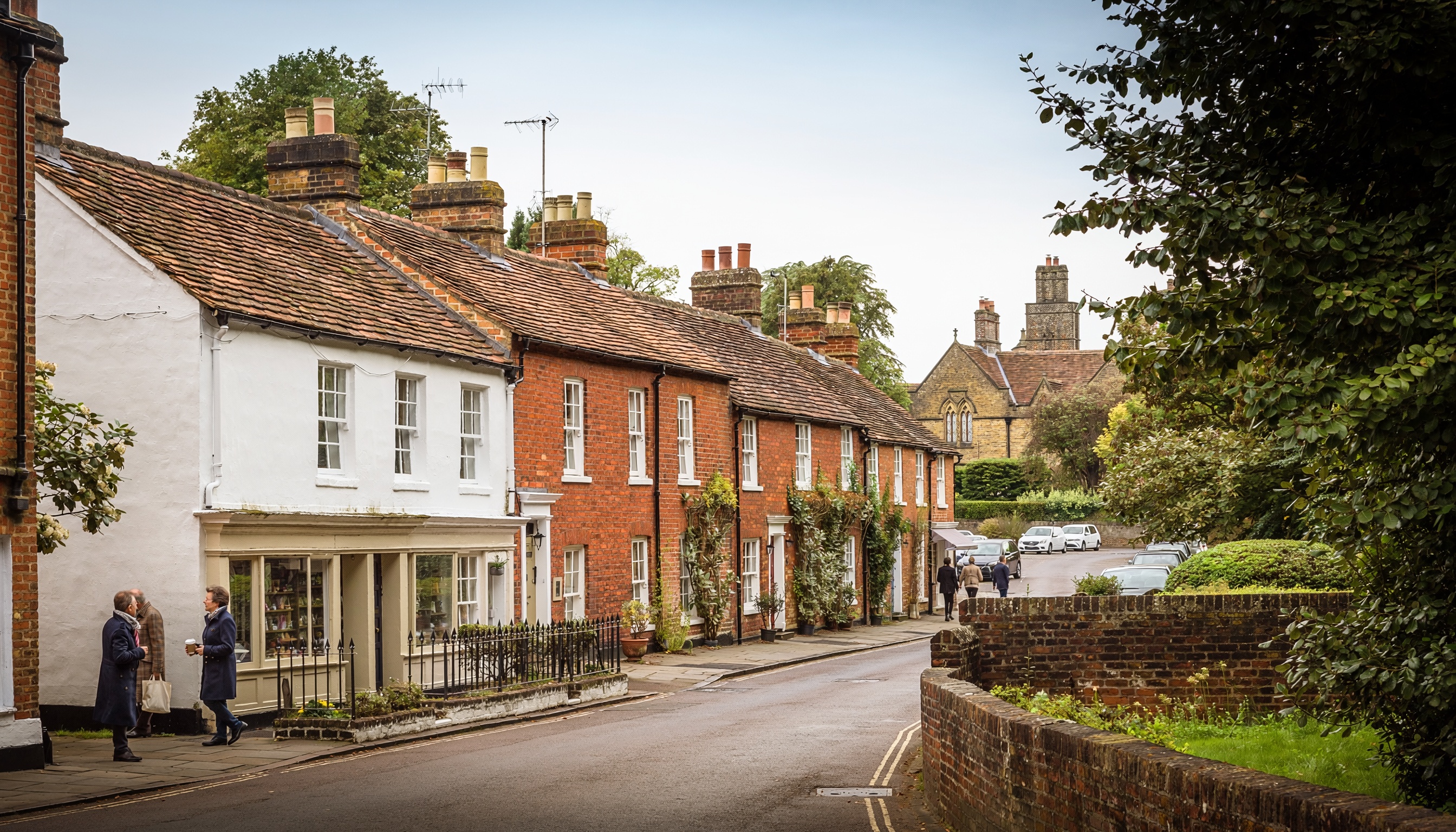 Charming village street with traditional brick and plaster houses, tiled roofs, climbing plants, parked cars, and a Gothic-style stone building in the background.