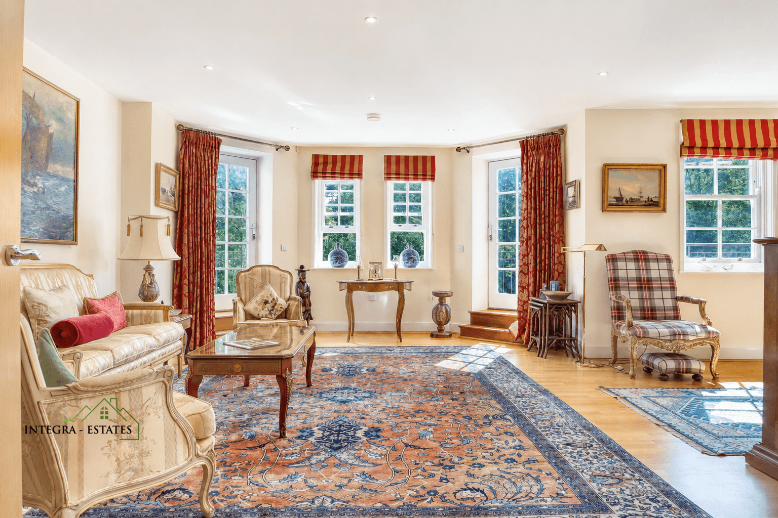 Elegant living room with cream walls, large windows framed by red patterned curtains, and vintage-style furniture including upholstered armchairs, a sofa with decorative cushions, and a wooden coffee table with glass top. A blue and orange ornate rug covers the wooden floor, complemented by framed paintings, vases, and a plaid armchair with footstool. Logo ‘INTEGRA ESTATES’ appears on the left side.