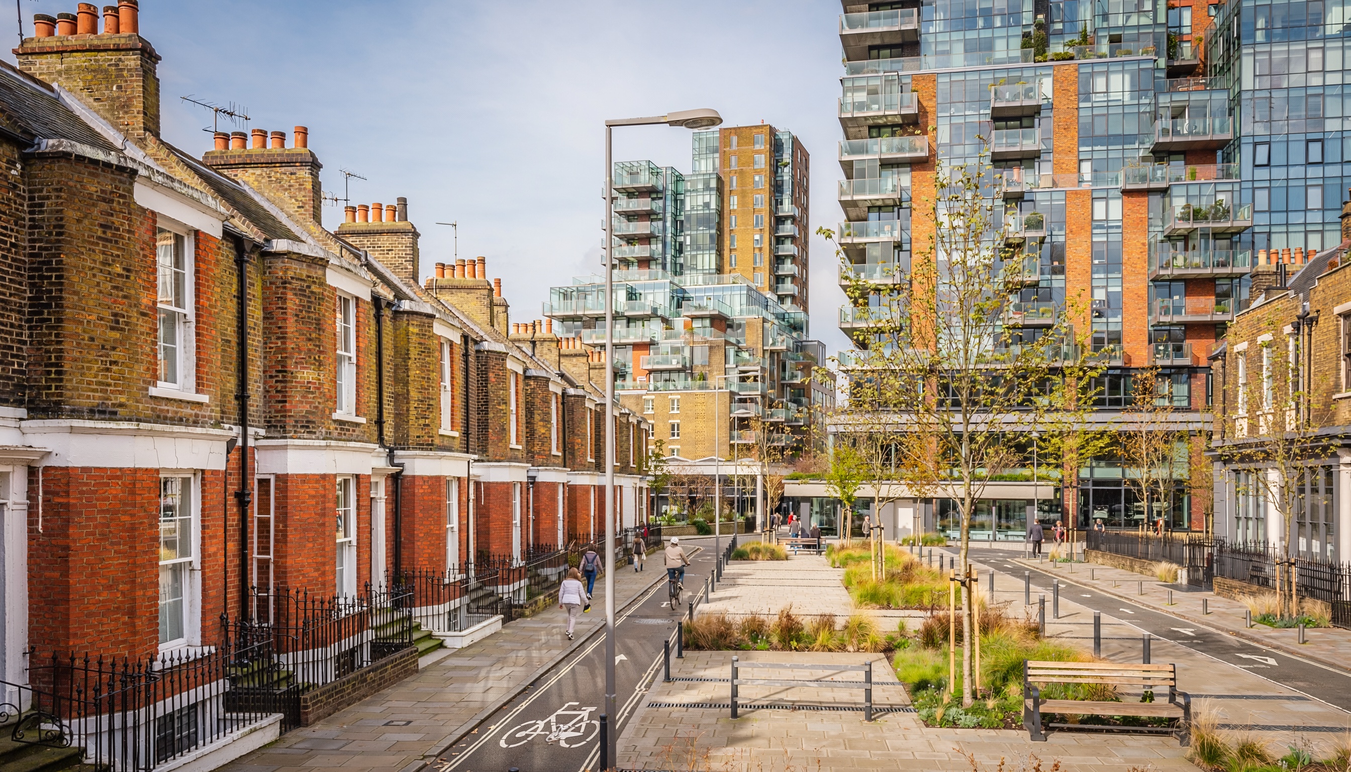 Urban street with rows of traditional brick terraced houses and chimneys in the foreground, contrasting with tall modern glass apartment buildings in the background, landscaped path with benches and people walking and cycling.