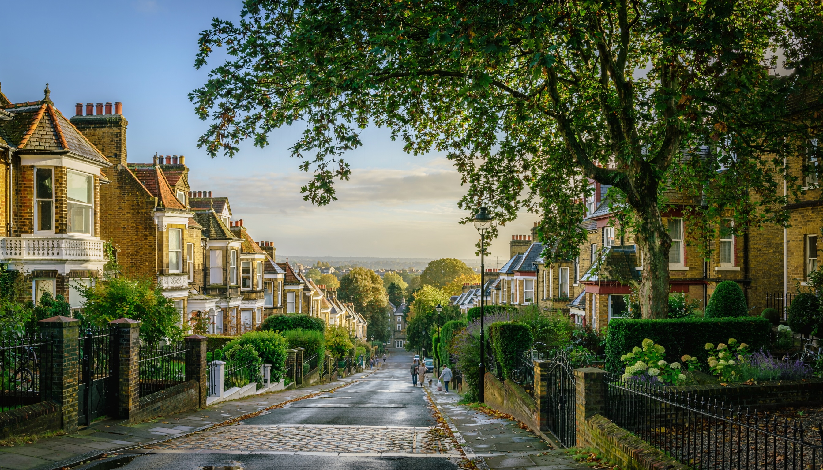 Picturesque residential street sloping downward, lined with traditional brick houses with bay windows and decorative trim, shaded by mature trees forming a canopy, with warm sunlight and people walking along the sidewalk.