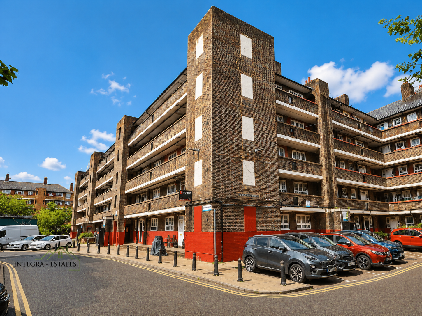 Multi-story residential building with brown brick exterior and red-painted lower sections, featuring balconies and rows of white-framed windows. Paved area in front with parked cars in gray, red, and white, separated from the street by bollards. Clear sky with scattered clouds above. Signs on the building read ‘ACORN’ and ‘INTEGRA ESTATES.