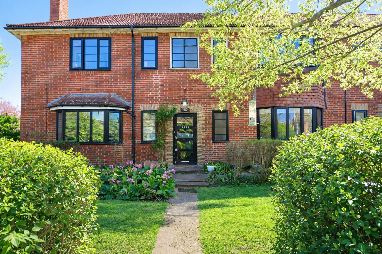 A two‑story brick house with a red‑tiled roof and black‑framed windows. The black front door, set within a decorative brick arch, is flanked by two bay windows. A paved walkway leads through a well‑kept garden of green grass, flowering plants, and trimmed bushes. Sunlight brightens the scene, with a tree partially framing the view from the right.