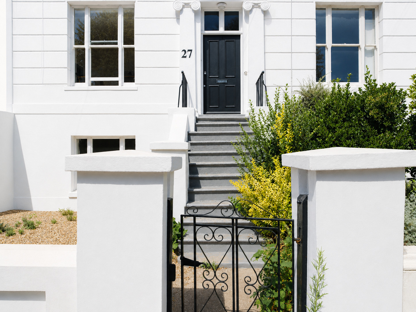 Front entrance of a white residential building with a black door marked number 27, elevated by gray steps with black railings and a small wrought-iron gate. The entryway is framed by white pillars, shrubs, and plants, with large rectangular windows above, combining modern cleanliness with classical detailing.