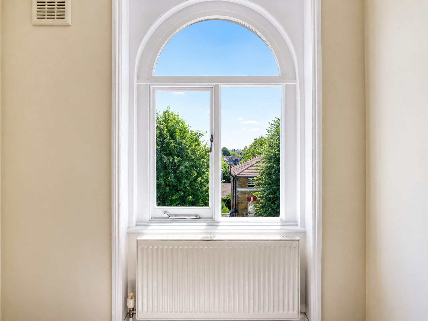 Interior view of a bright room with a large arched window framed in white, a radiator beneath it, and a view of green trees and houses under a blue sky. Natural light highlights the clean, minimal design and symmetry, connecting the indoor space with the outdoors