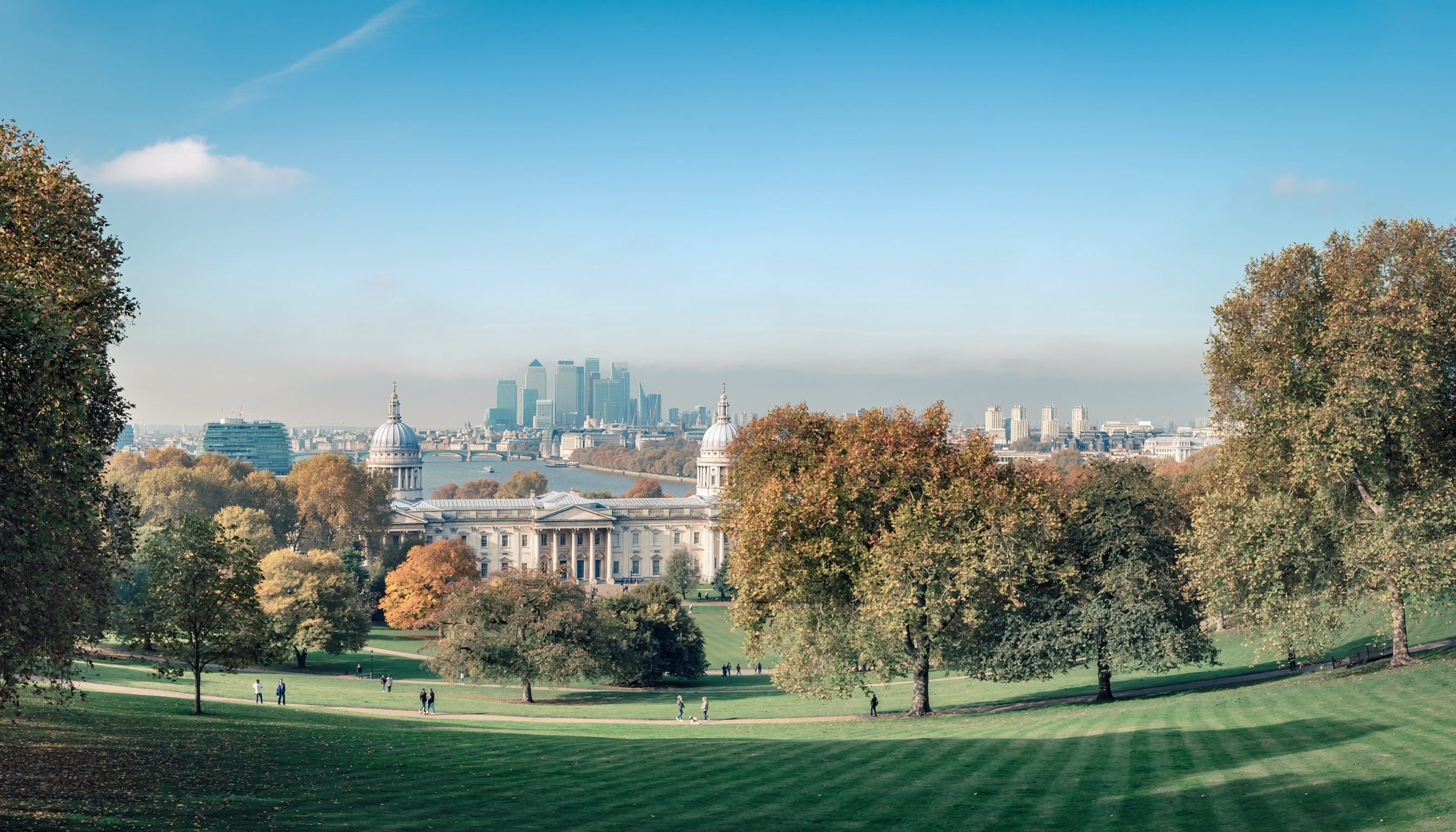 Scenic view of Greenwich Park with green lawns and autumn trees, the Old Royal Naval College with domed towers, River Thames beyond, and Canary Wharf skyscrapers rising against a clear blue sky.