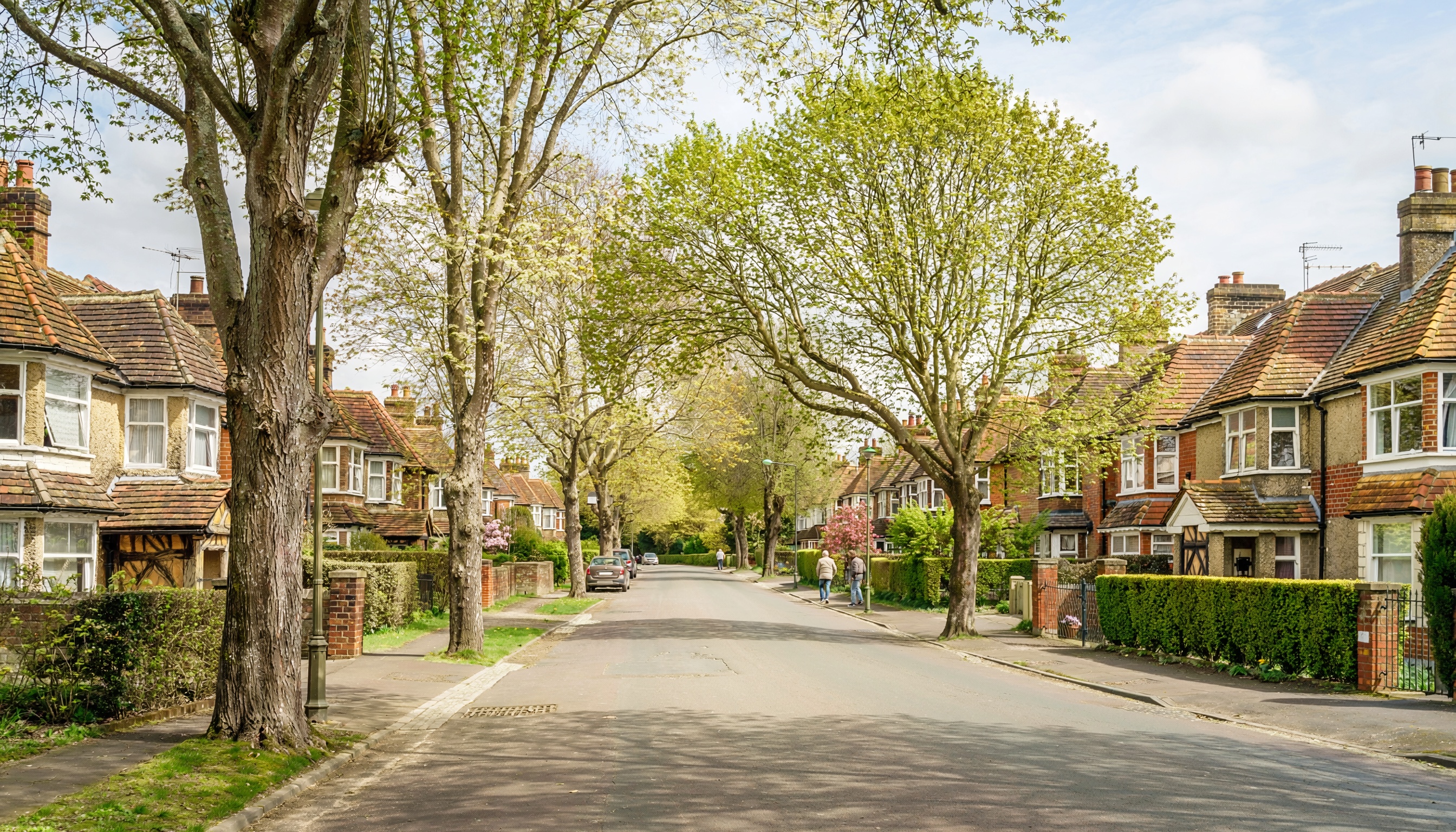 Quiet suburban street with semi-detached brick and stucco houses, pitched tiled roofs, leafy canopy of mature trees, low hedges and brick walls, parked cars, and people walking along the sidewalks in springtime.
