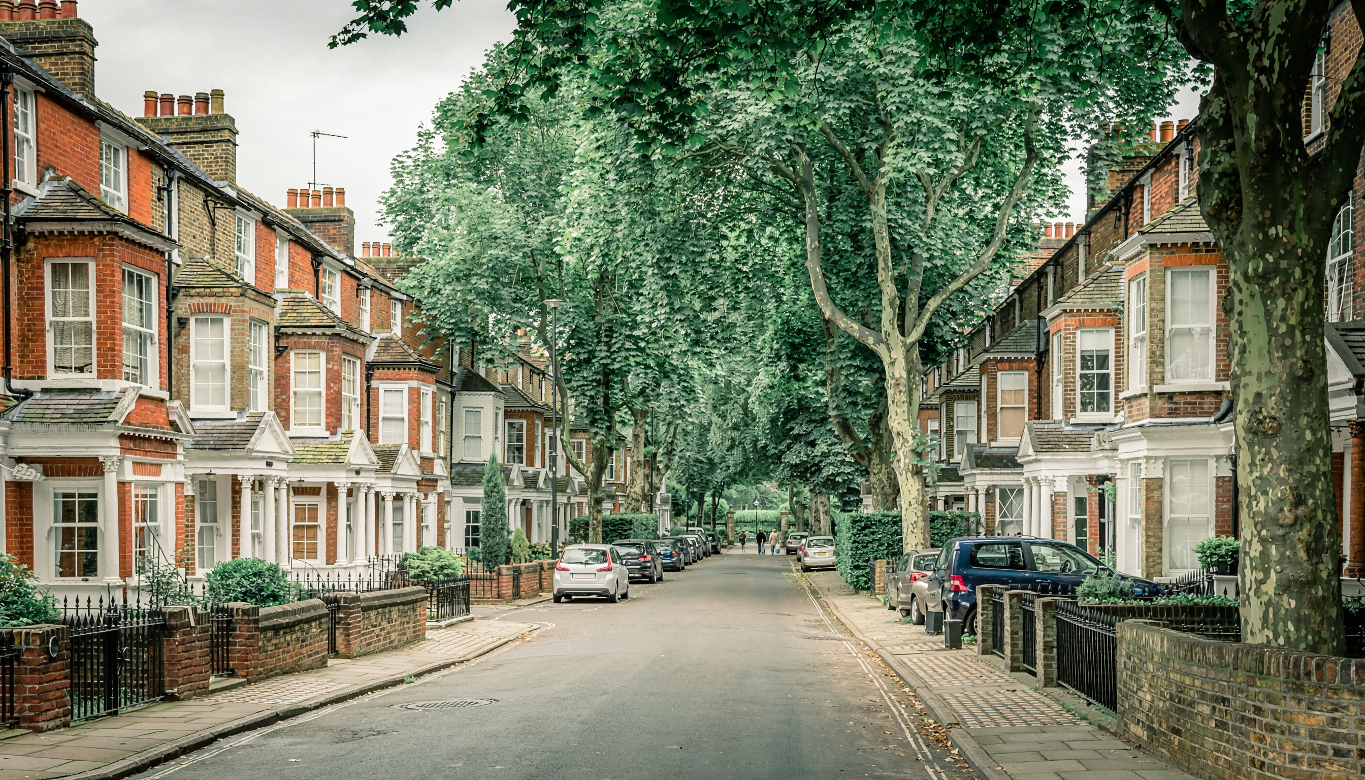 “Residential street lined with Victorian-style brick houses, bay windows and white trim, shaded by mature trees forming a leafy tunnel, with small front gardens, parked cars, and people walking in the distance.