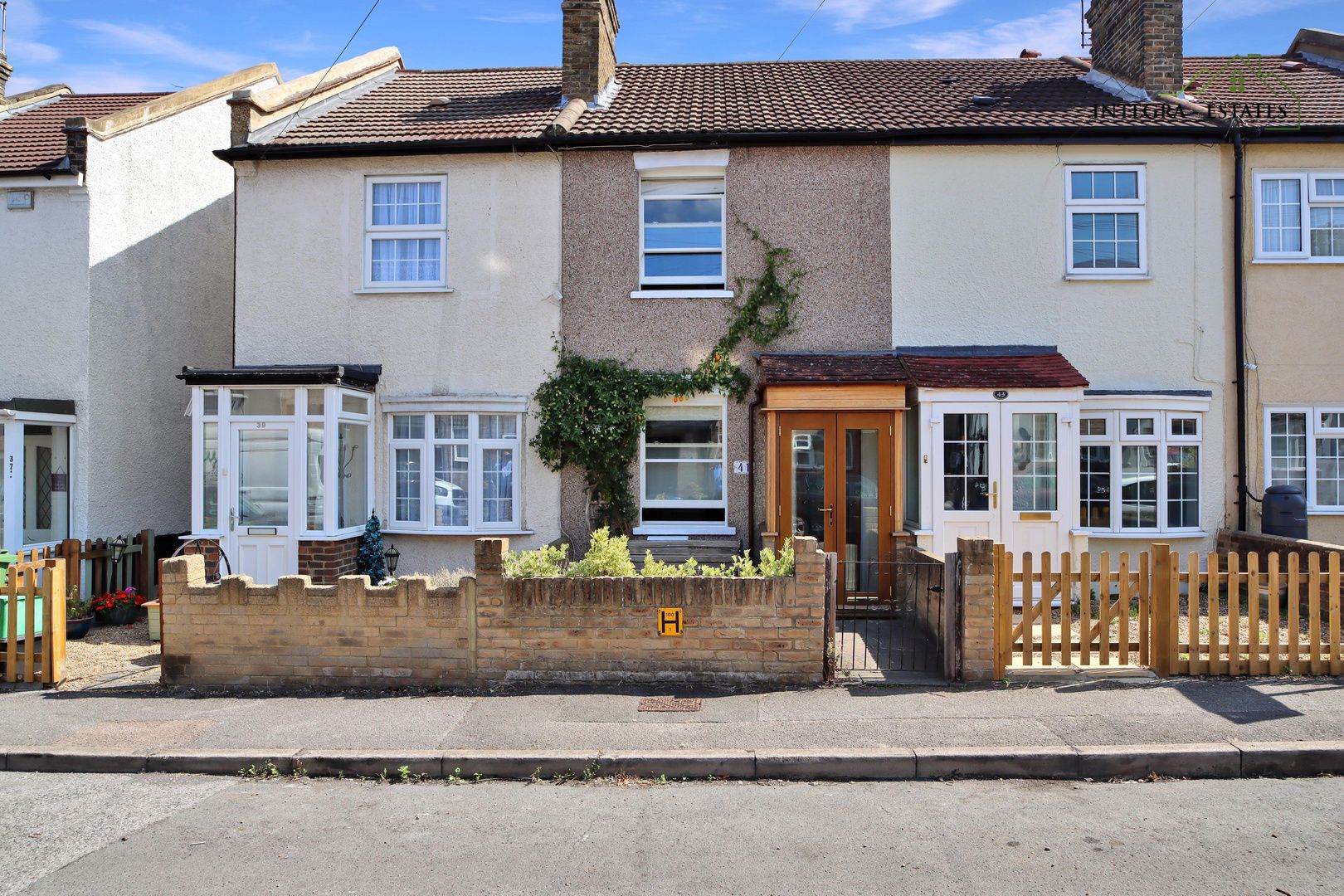 A charming mid-terraced house with a small front garden bordered by a low brick wall, featuring a mix of cream and pebble-dash exterior, white-framed windows, and a wooden porch entrance under a tiled roof on a quiet residential street.
