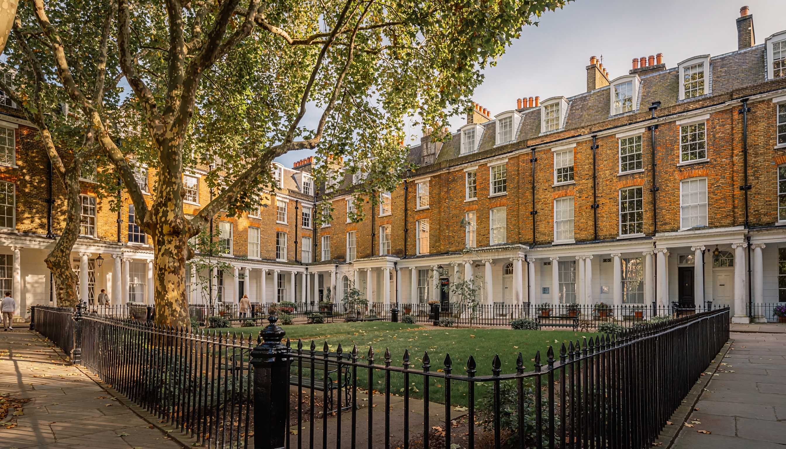 Georgian-style brick townhouses with tall sash windows, white porticos, and chimneys surrounding a courtyard with grassy area, benches, and mature trees in bright sunlight.