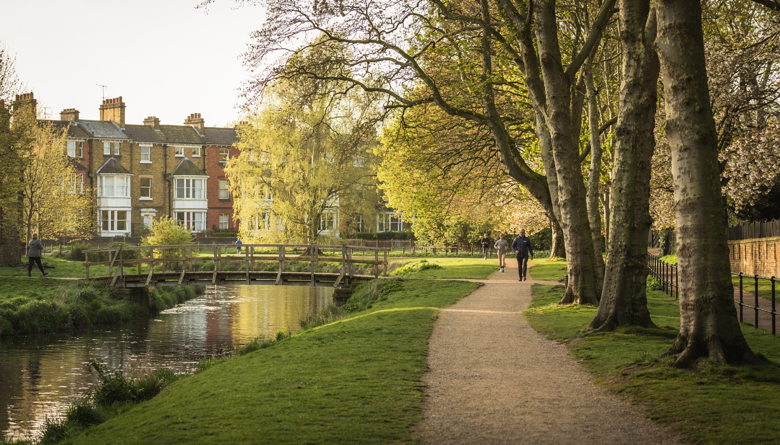 Scenic riverside path with tall spring trees, calm river and wooden footbridge, multi-story brick houses in the background, and people walking in warm late afternoon sunlight.