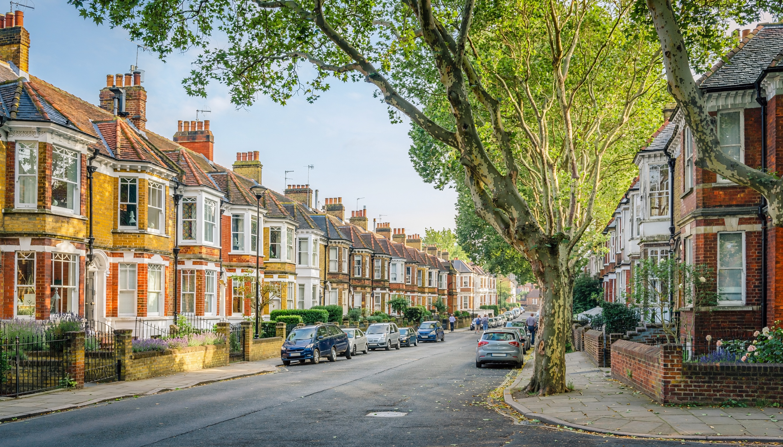 Residential street lined with Victorian terraced houses featuring brick facades, bay windows, tiled roofs and chimneys, shaded by mature green trees forming a canopy, with cars parked along both sides in daytime.