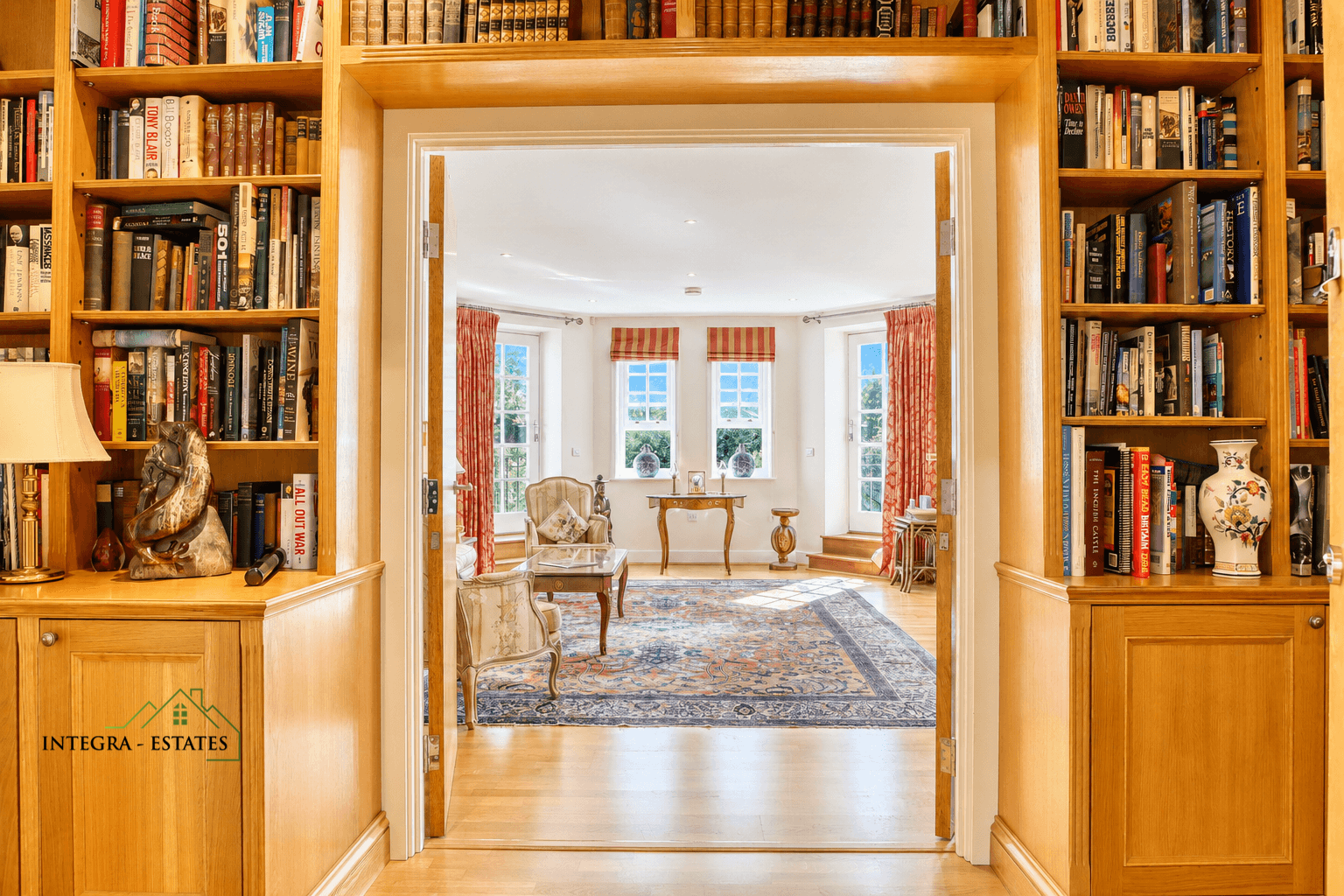 Interior view from a library with built-in light wood bookshelves filled with books, decorative items, and a lamp, looking into a bright sitting room. The sitting room features large windows with red and orange striped curtains, classic armchairs, a wooden table, and a patterned rug on polished wooden floors. Logo ‘INTEGRA ESTATES’ appears on the lower left cabinet.