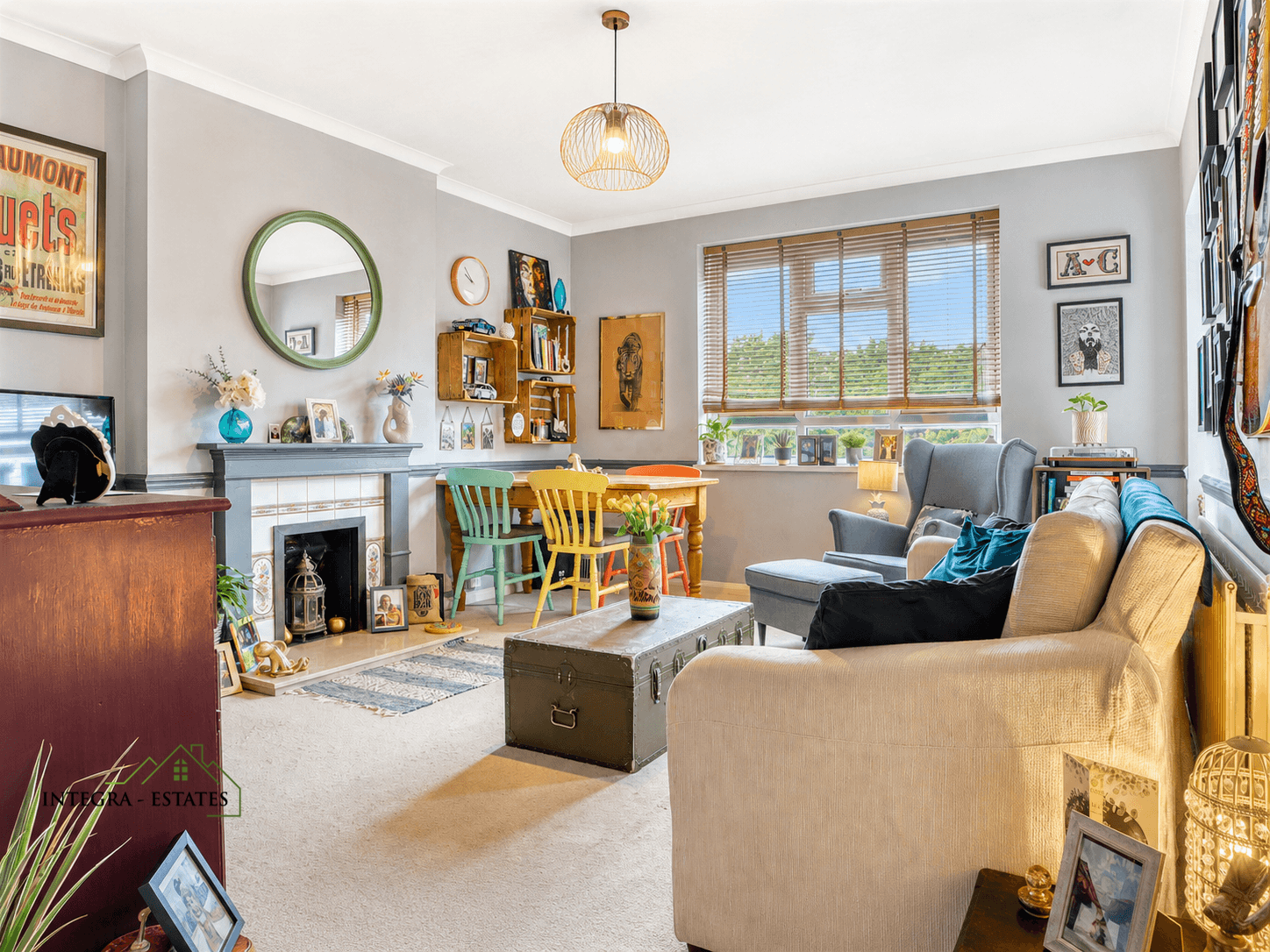 Cozy living and dining area with eclectic mix of vintage and modern decor. Light gray walls, beige carpet, and large window with wooden blinds. Beige sofa with blue and black cushions, gray armchair, and dark green trunk as coffee table. Dining area with wooden table and mismatched green and yellow chairs. Fireplace with gray mantel, decorative tiles, vases, flowers, and round green-framed mirror above. Wooden crate shelves holding books and decor, framed artworks and photographs on walls.