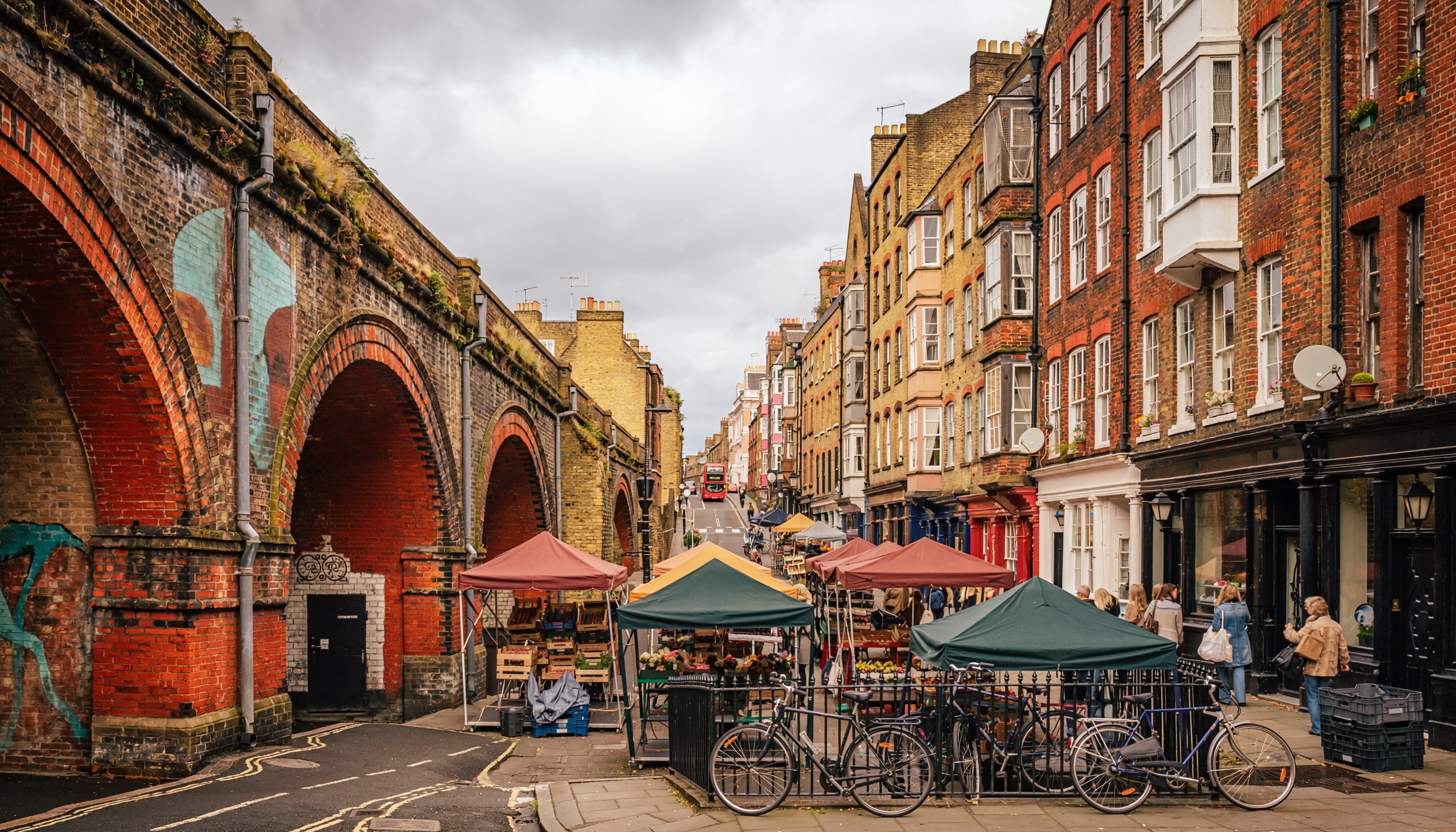Lively urban street market with colorful canopies set up between historic brick buildings and a railway viaduct with painted arches, bicycles parked in foreground, and a red double-decker bus visible in the distance.