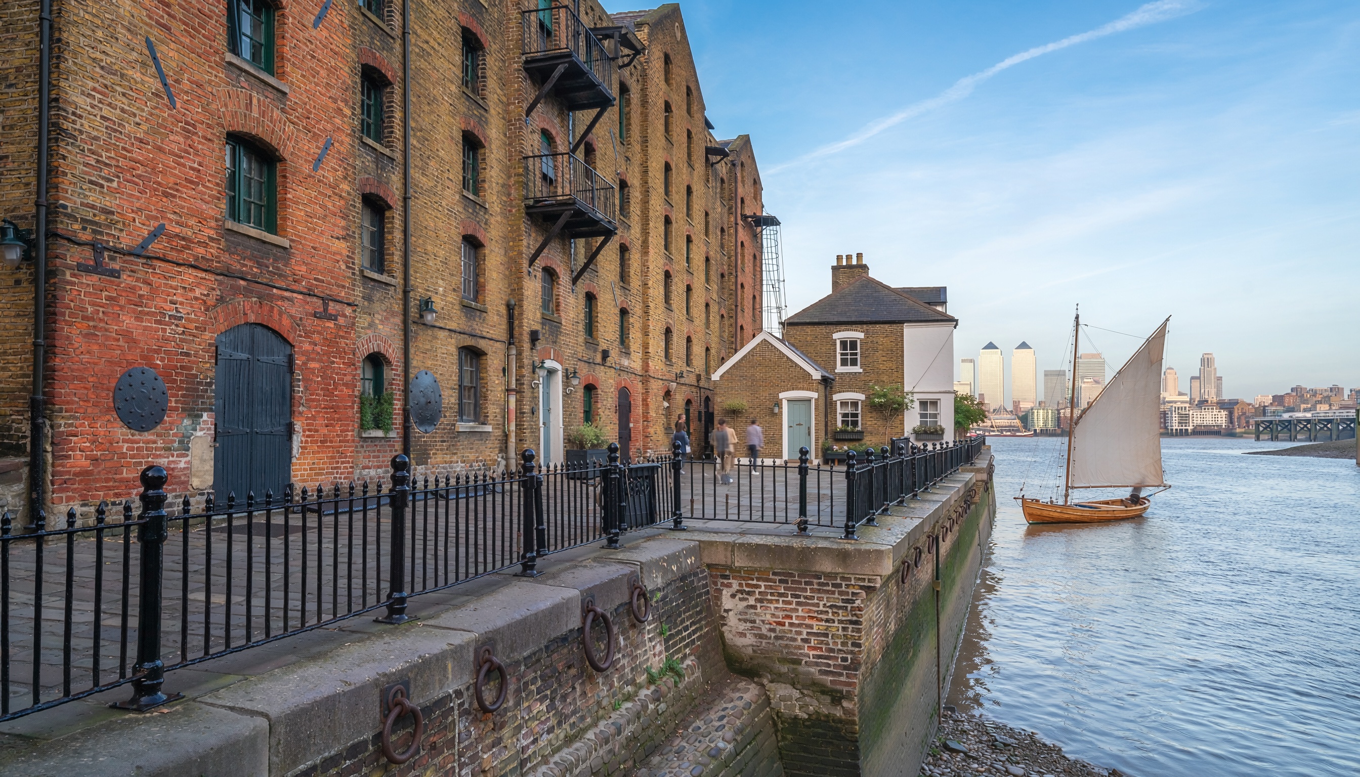 Riverside scene with historic brick warehouses featuring arched windows and iron railings along a cobblestone quay, a small sailboat on the river, and modern skyscrapers across the water in contrast.