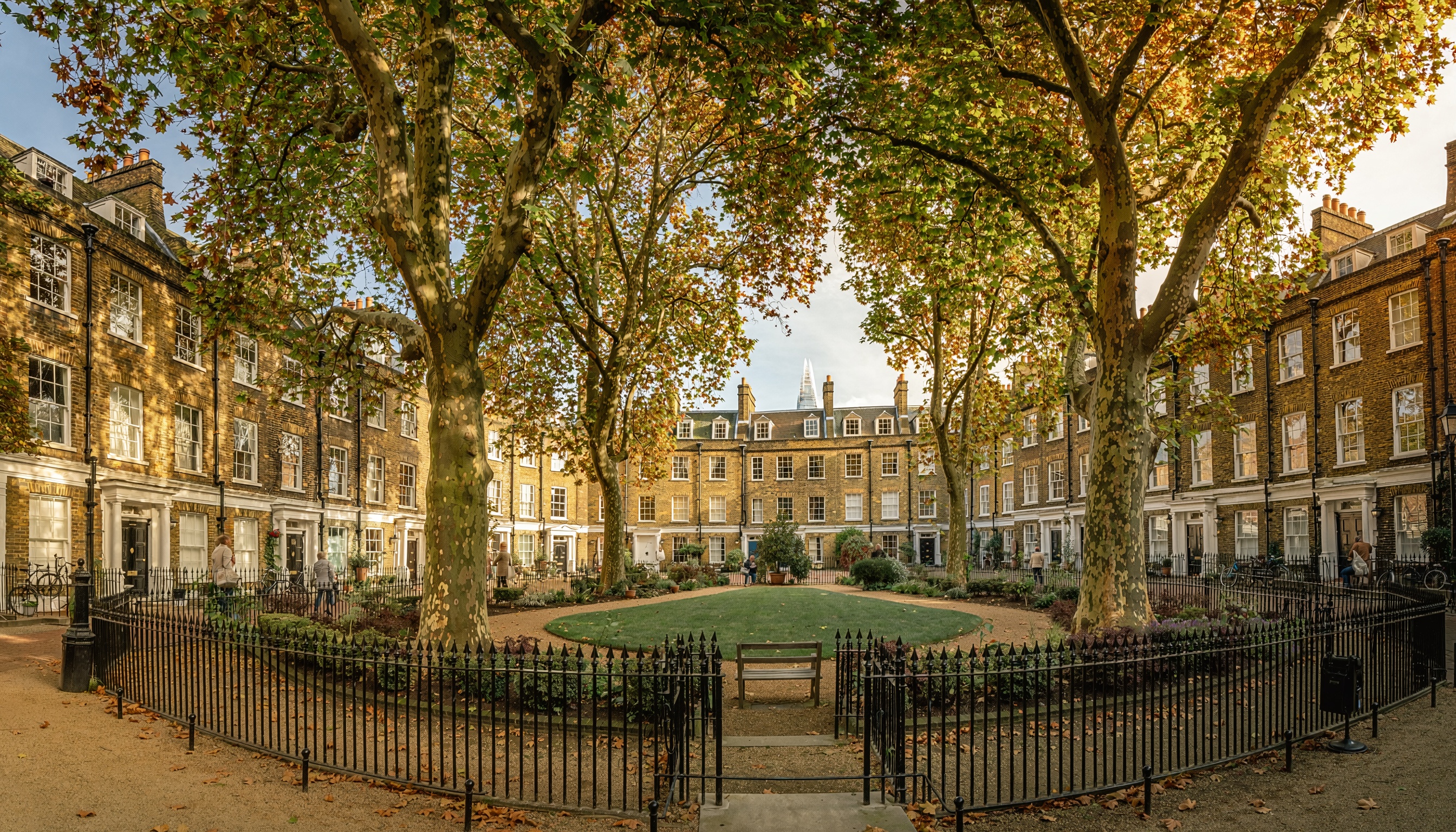 Georgian-style residential square with rows of brick townhouses featuring white-trimmed windows and dormer roofs, surrounding a central park with circular lawn, benches, and mature autumn trees enclosed by wrought-iron fencing.