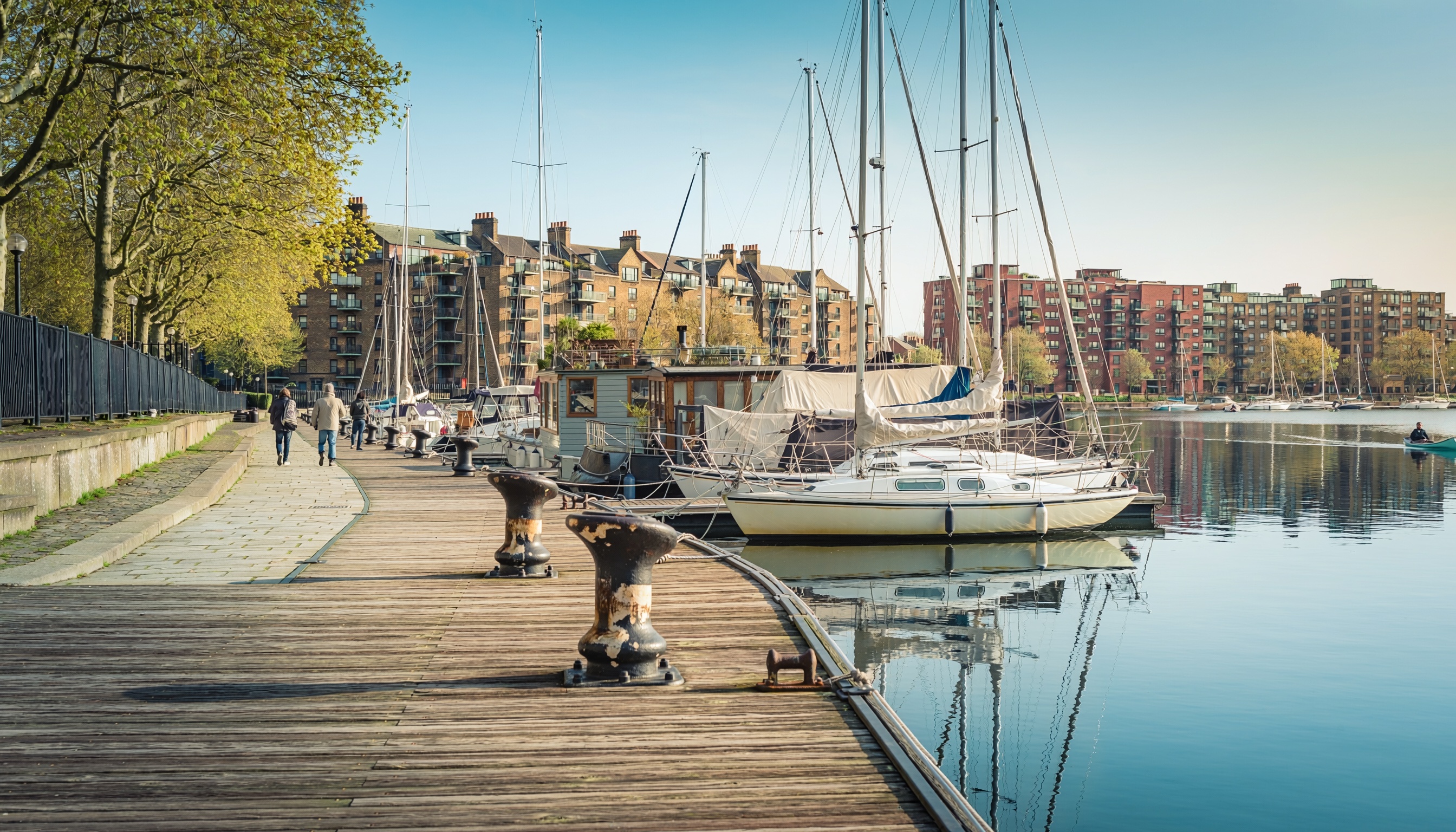 Peaceful marina with sailboats and yachts docked along a wooden pier, people walking on a tree-lined promenade with black metal fence, multi-story brick residential buildings with balconies in the background, and calm water reflecting boats and buildings under a clear blue sky.