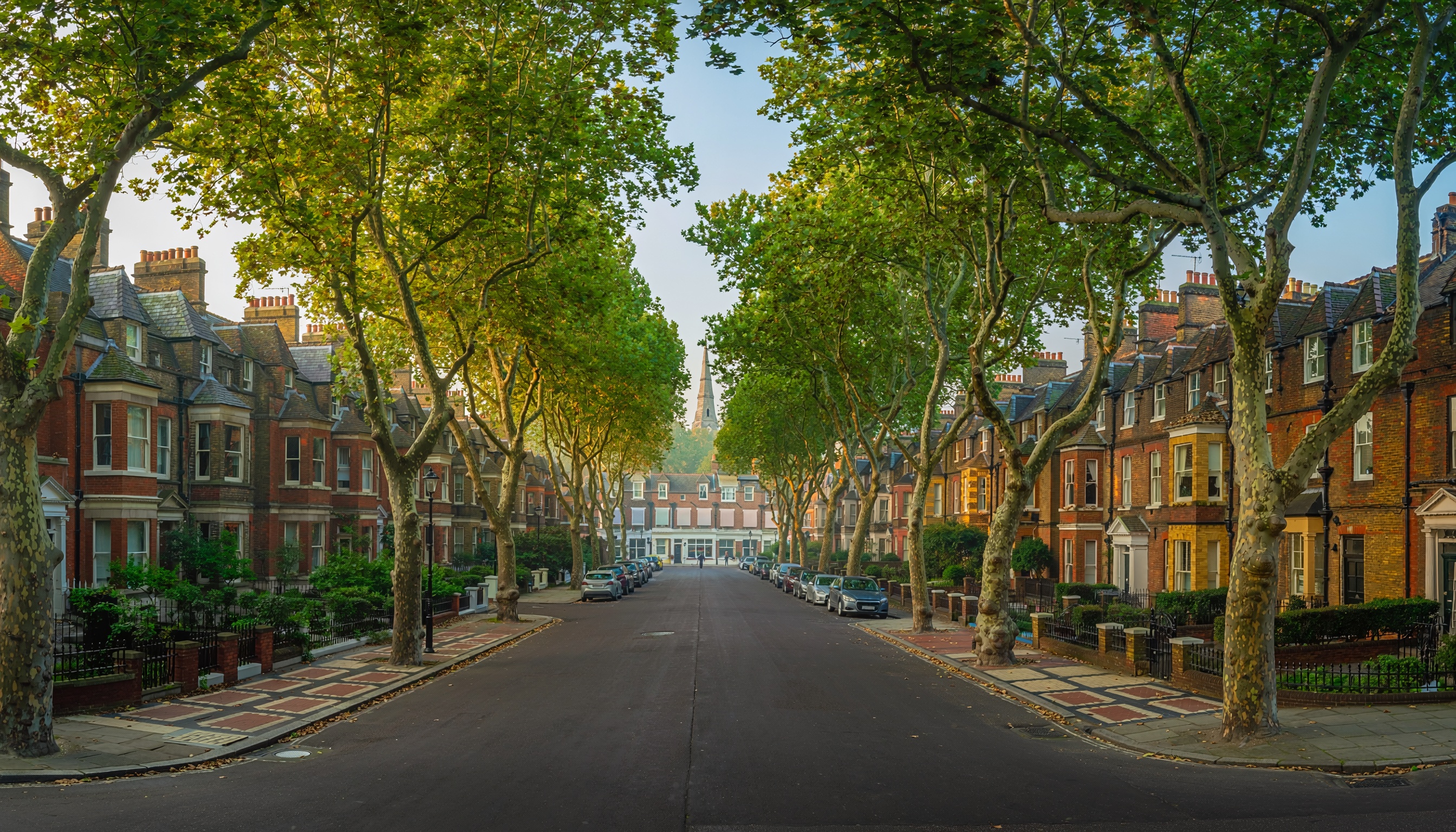 Picturesque residential street lined with tall mature trees forming a green canopy, rows of late 19th-century brick townhouses with slate roofs, chimneys, and large windows, patterned pavements and iron railings, with a larger building and church spire visible in the distance.