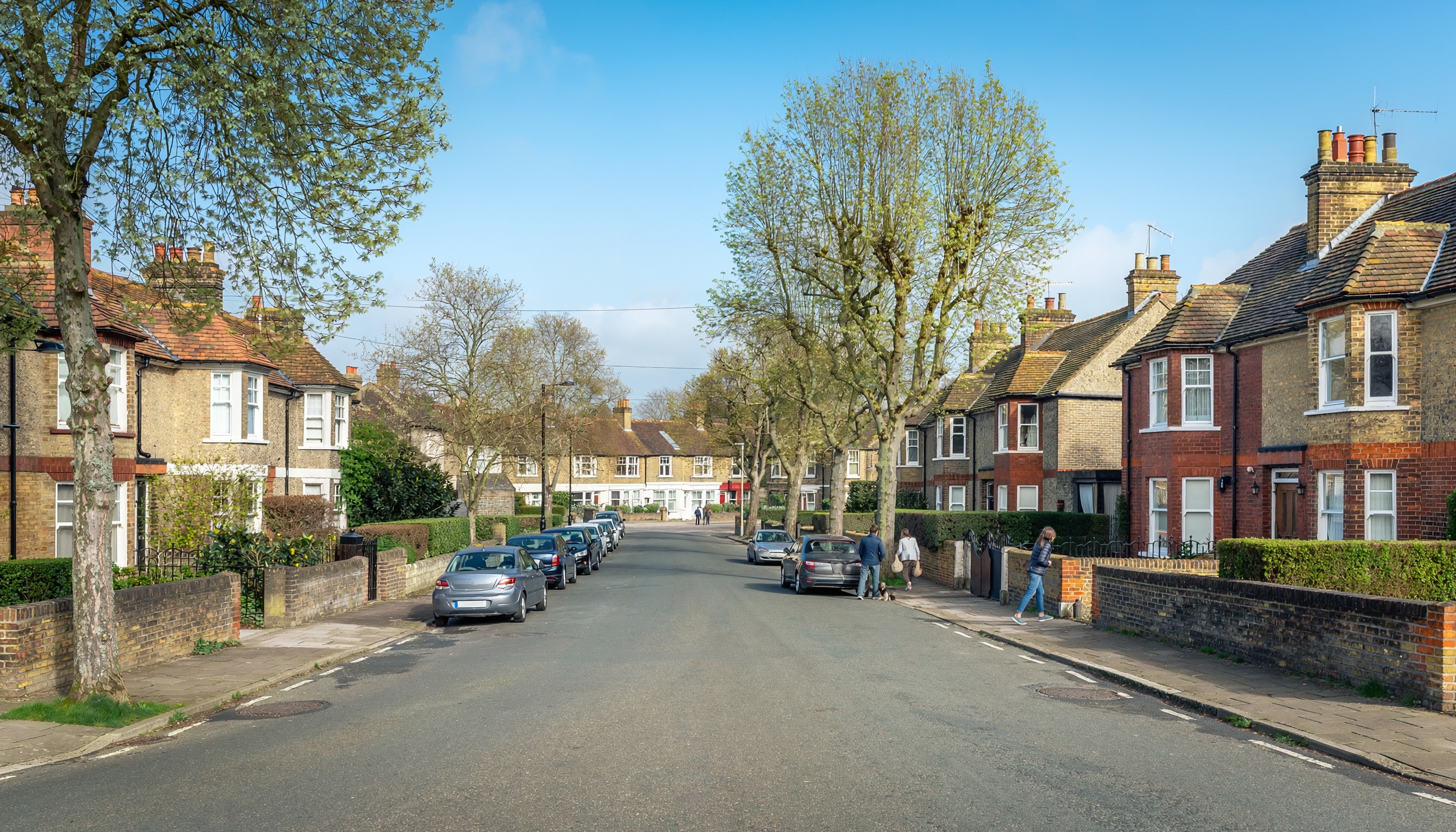 Quiet residential street with rows of brick houses featuring tiled roofs and white-framed windows, small front gardens with hedges and shrubs, cars parked along the curb, and people walking under leafy spring trees on a clear day.