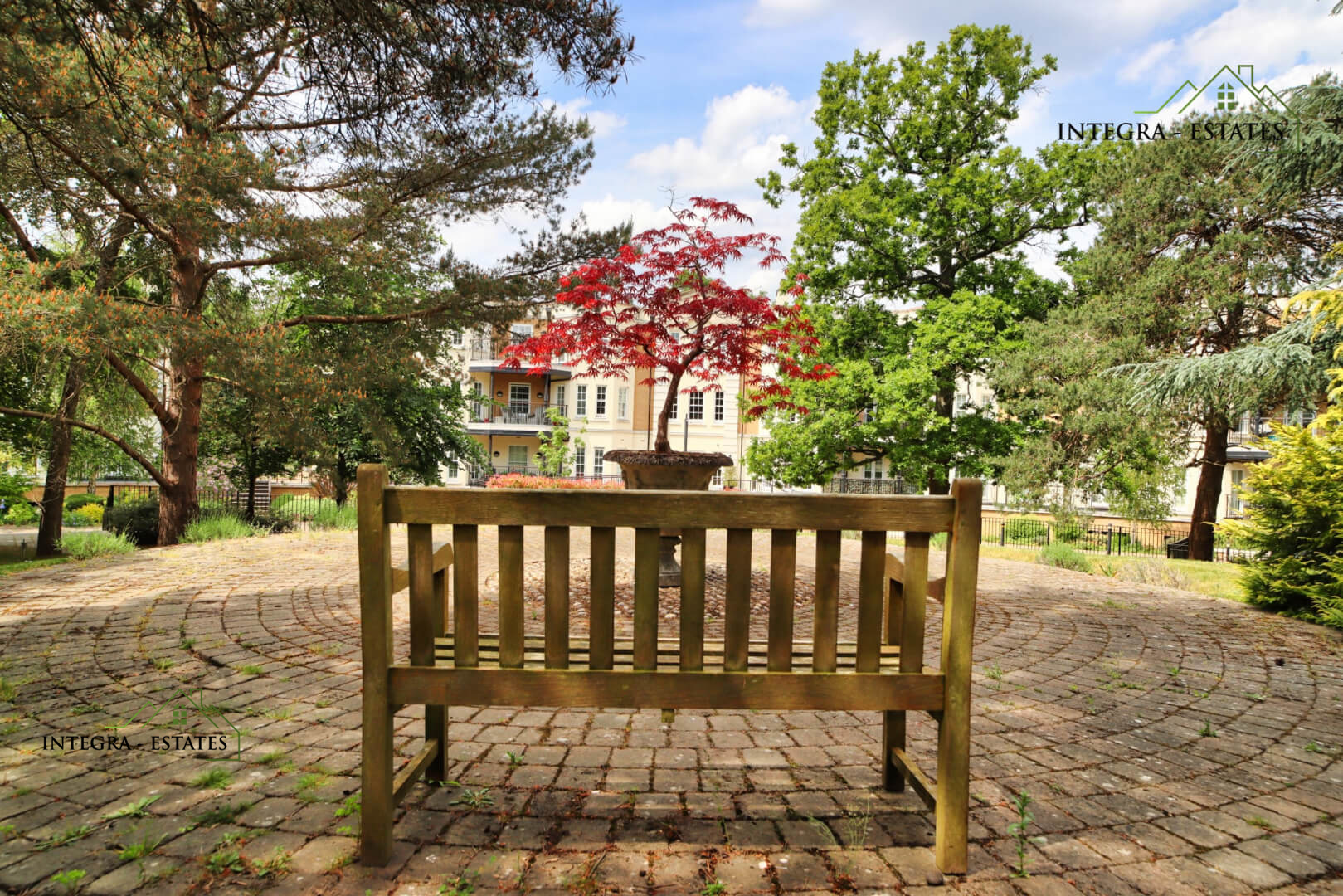Wooden bench on a circular brick-paved area with a planter holding a small Japanese maple tree with bright red leaves. Surrounded by lush greenery and residential buildings with balconies and white facades, under a partly cloudy sky. Logo ‘INTEGRA ESTATES’ with stylized house graphic appears in the corners.”