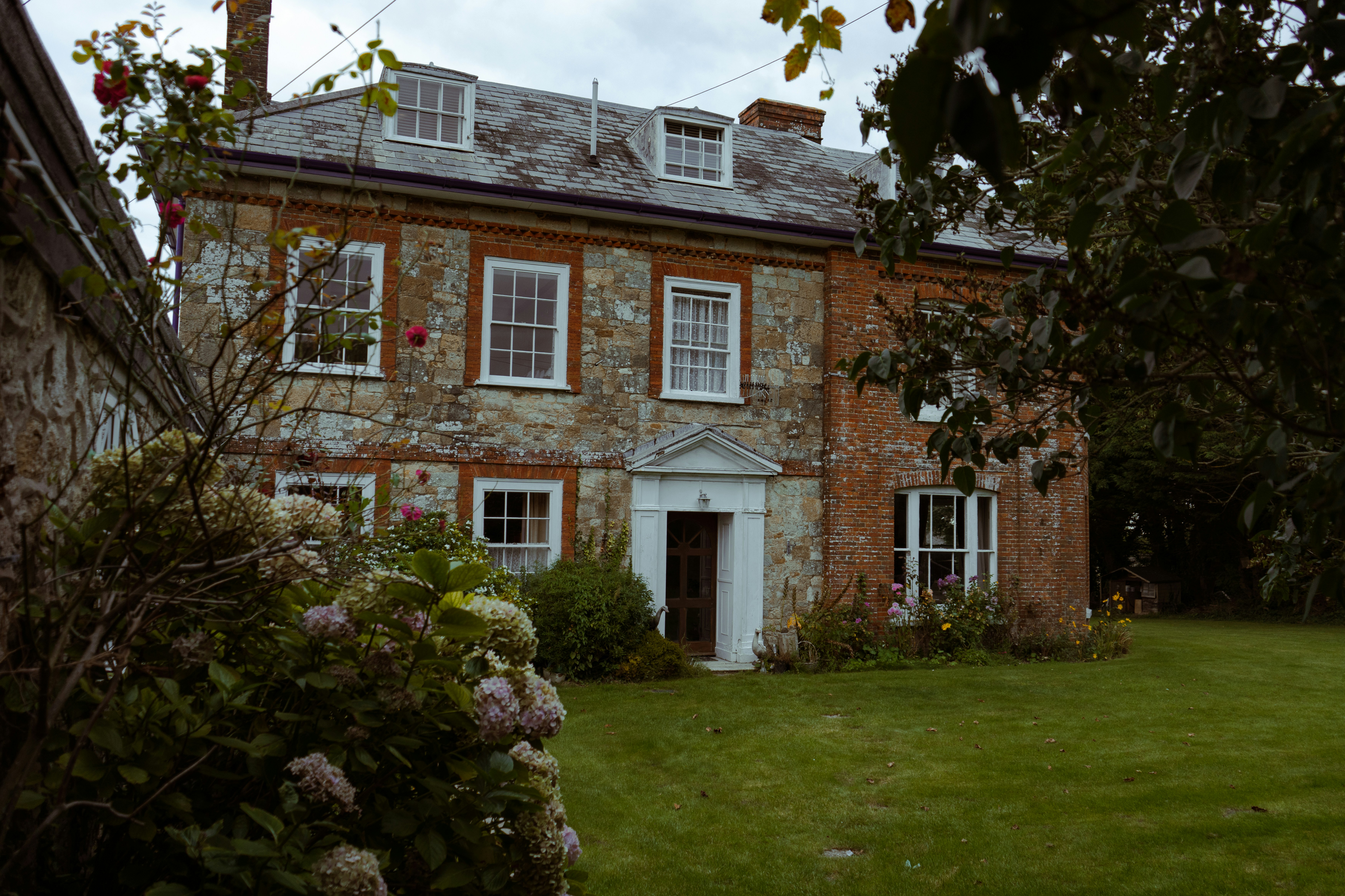 A traditional two-story house with stone and brick exterior walls, white-framed windows, and a slate roof with dormer windows. A central front door with a columned portico anchors the facade. The home is surrounded by a lush garden with flowering plants and a manicured lawn, evoking rustic charm and serenity.