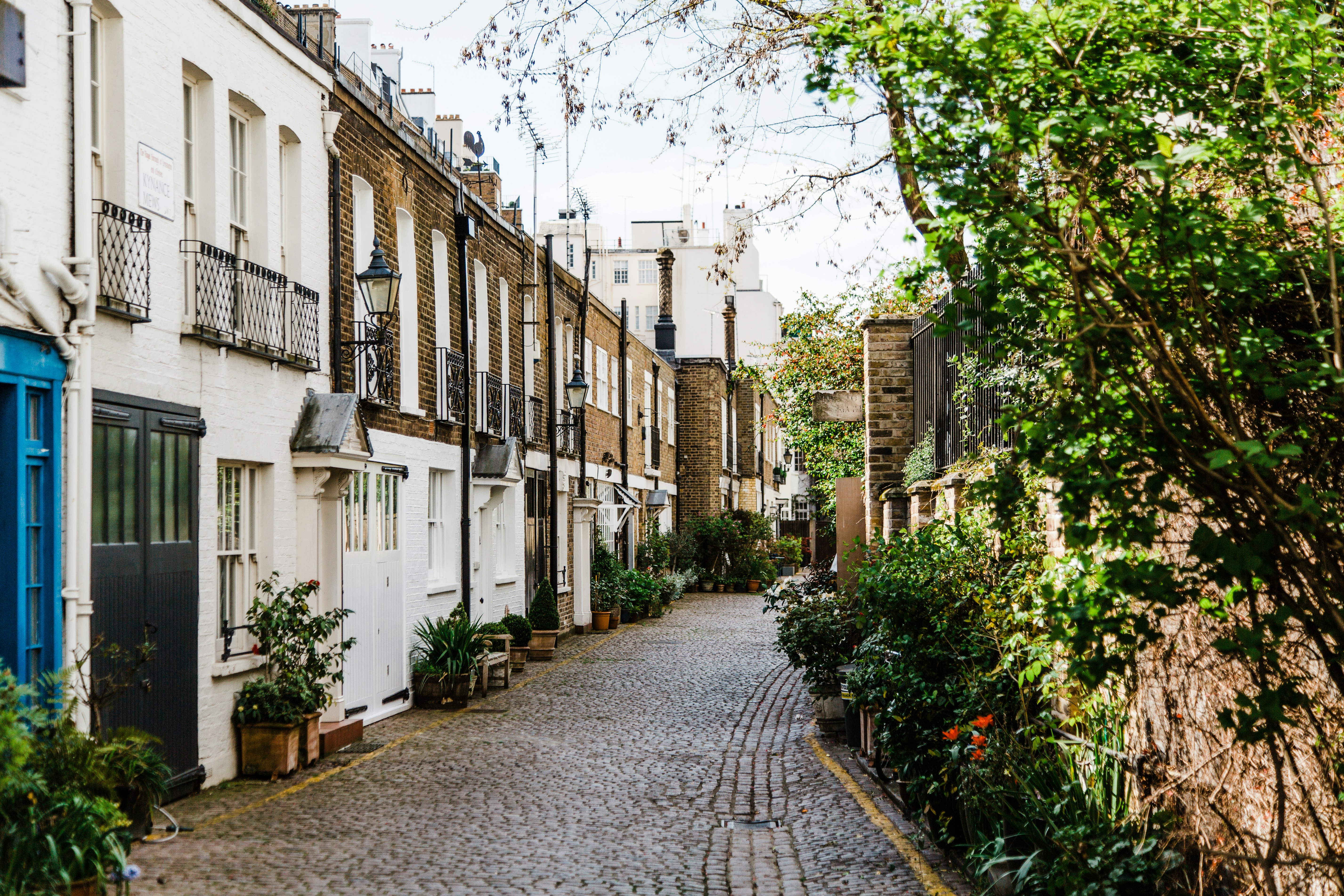 A narrow cobblestone street lined with traditional terraced houses, featuring brick and white-painted facades, wrought iron balconies, and classic street lamps. Potted plants and greenery soften the urban setting, creating a peaceful, picturesque atmosphere suggestive of a historic European neighborhood.