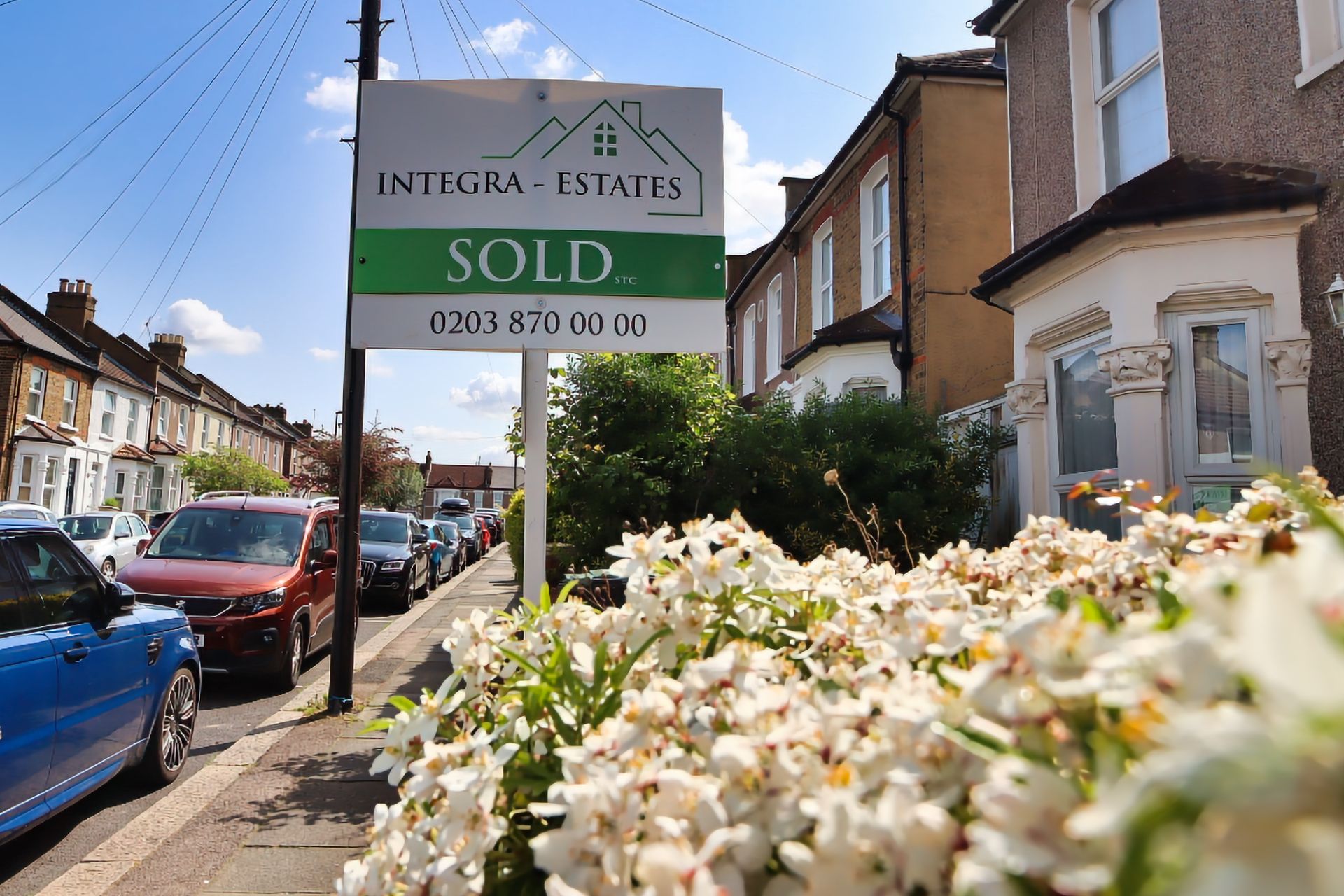 A “SOLD” sign from Integra Estates stands on a sunny residential street lined with terraced houses and parked cars. The sign is positioned in the foreground next to a patch of white flowers, while the background shows a row of traditional London-style homes, blue sky, and scattered clouds.