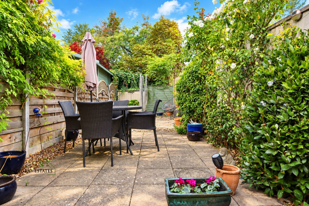 Private rear garden with a paved patio, outdoor dining set and parasol, surrounded by mature shrubs, climbing plants, and trees under a bright blue sky.
