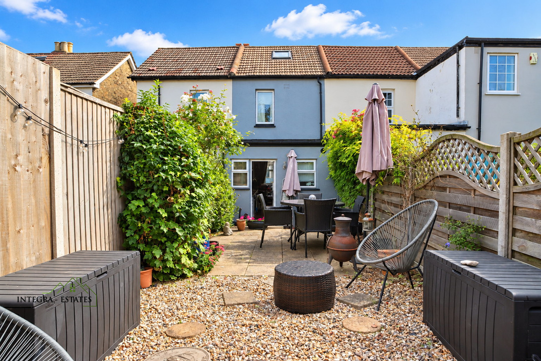 View from the rear garden toward a two-storey terraced home, featuring a gravel seating area, patio dining set with parasols, mature planting, and fenced boundaries under a blue sky.