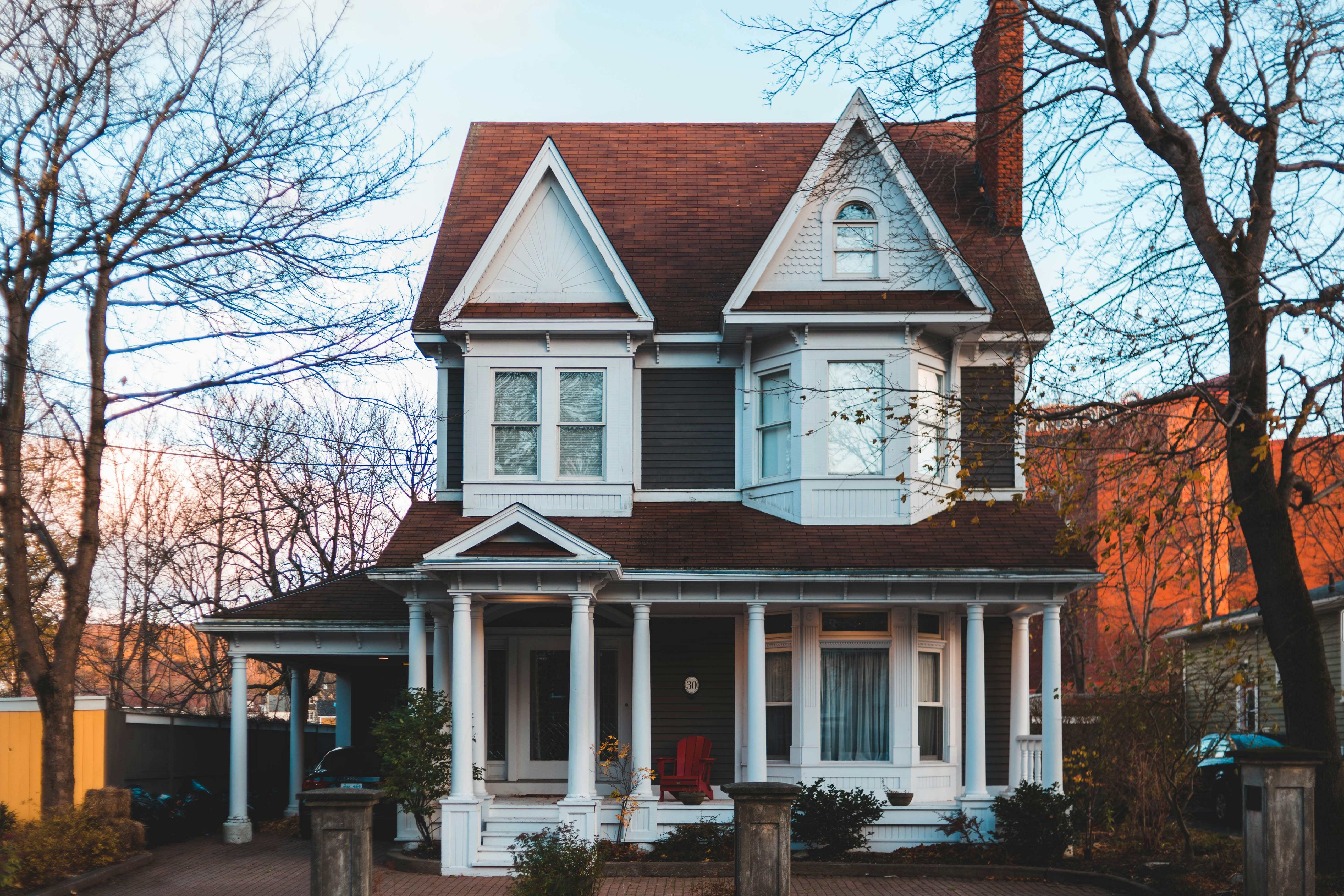 A two-story Victorian-style house features a steeply pitched roof, gables, and a prominent front porch with white columns. The exterior combines dark gray siding with white trim and decorative woodwork. A red chair sits on the porch, and a car is parked under a carport to the left. Leafless trees surround the home, suggesting late autumn or early winter.