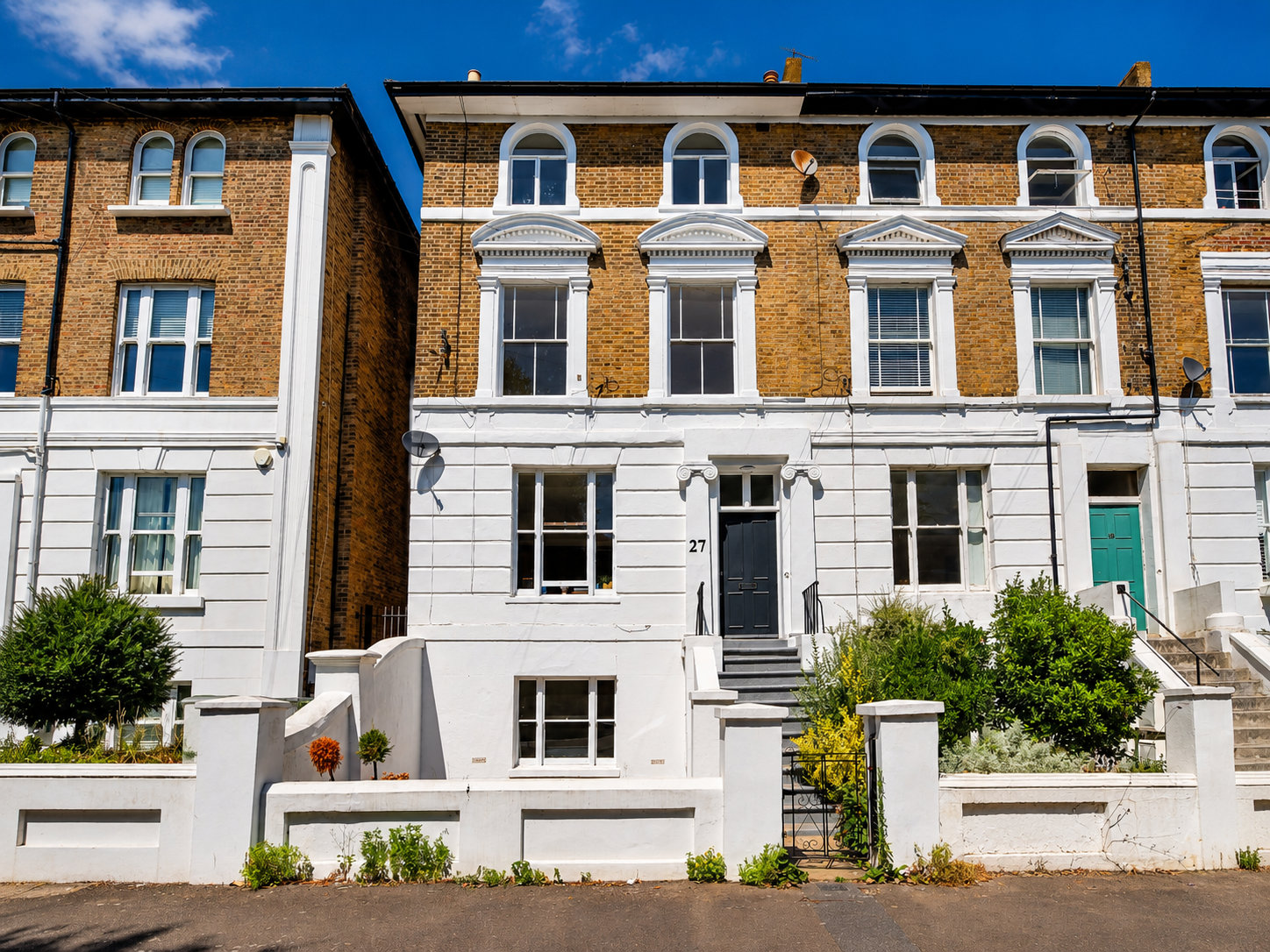 Row of traditional terraced houses with brick upper façades and white-painted lower sections, featuring tall sash windows, decorative pediments, and arched top-floor windows. House number 27 stands out with a dark gray front door, short stairway with black railings, and a small front garden enclosed by a low white wall.