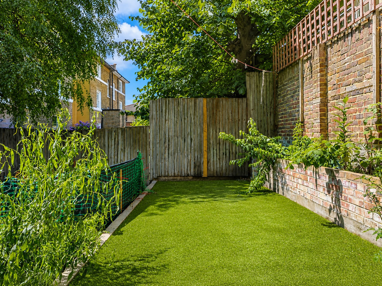 Small enclosed backyard with artificial grass, bordered by wooden fencing and a brick wall. Green plants and shrubs line the edges, a large tree provides shade, and a rope with clothespins is strung across the space. Residential brick buildings with white trim are visible in the background under a sunny blue sky.